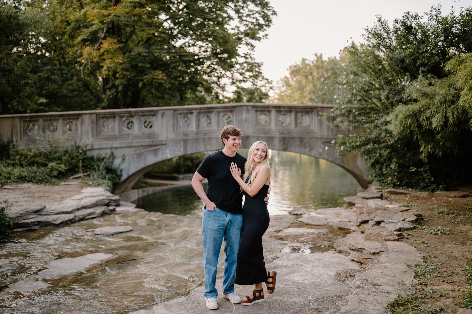 Couple posing together near a stone bridge over a creek, surrounded by lush trees in a scenic outdoor setting.