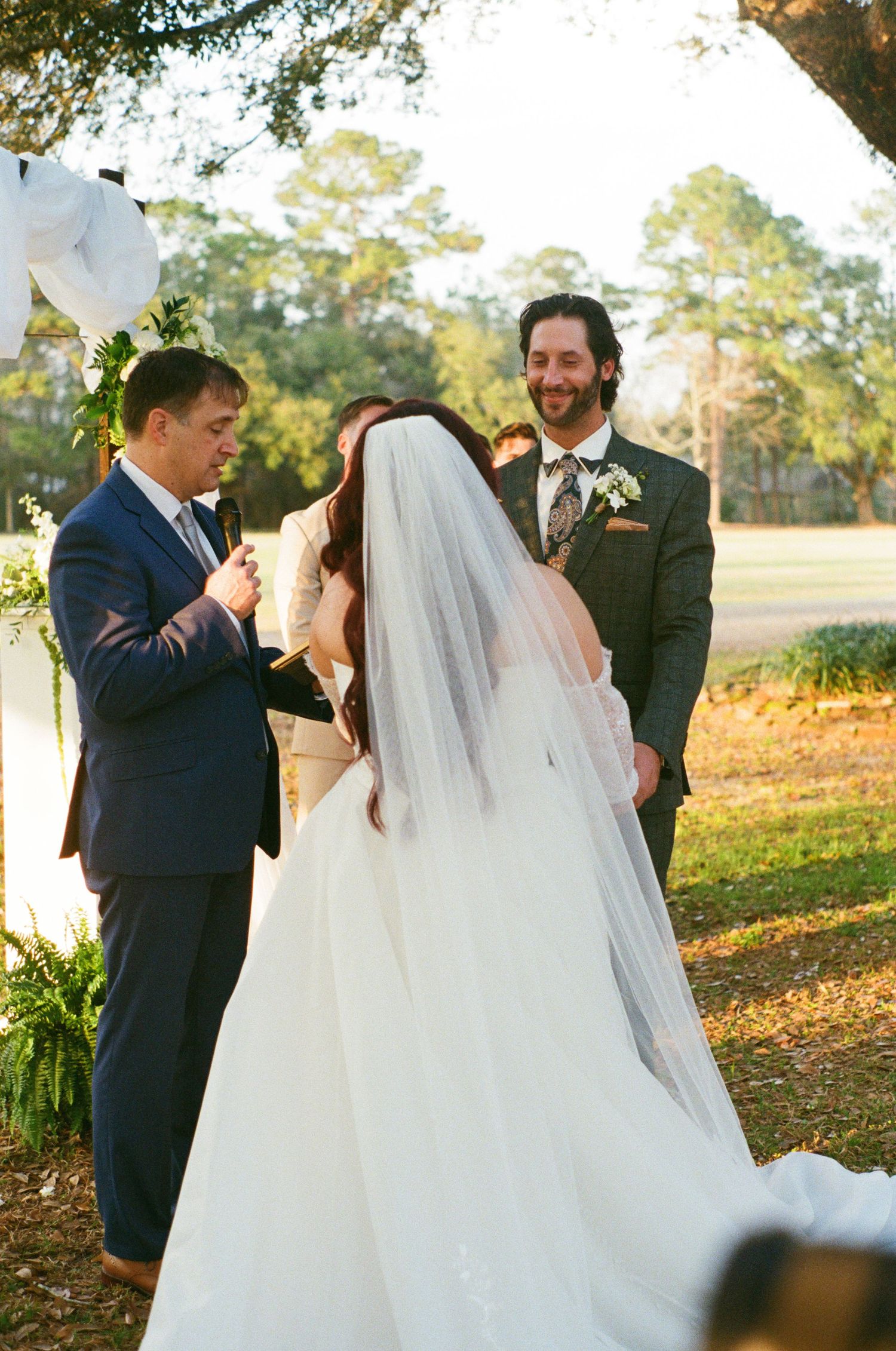 Bride in white gown with long veil holds groom's hand outdoors under red-leafed trees on their wedding day.