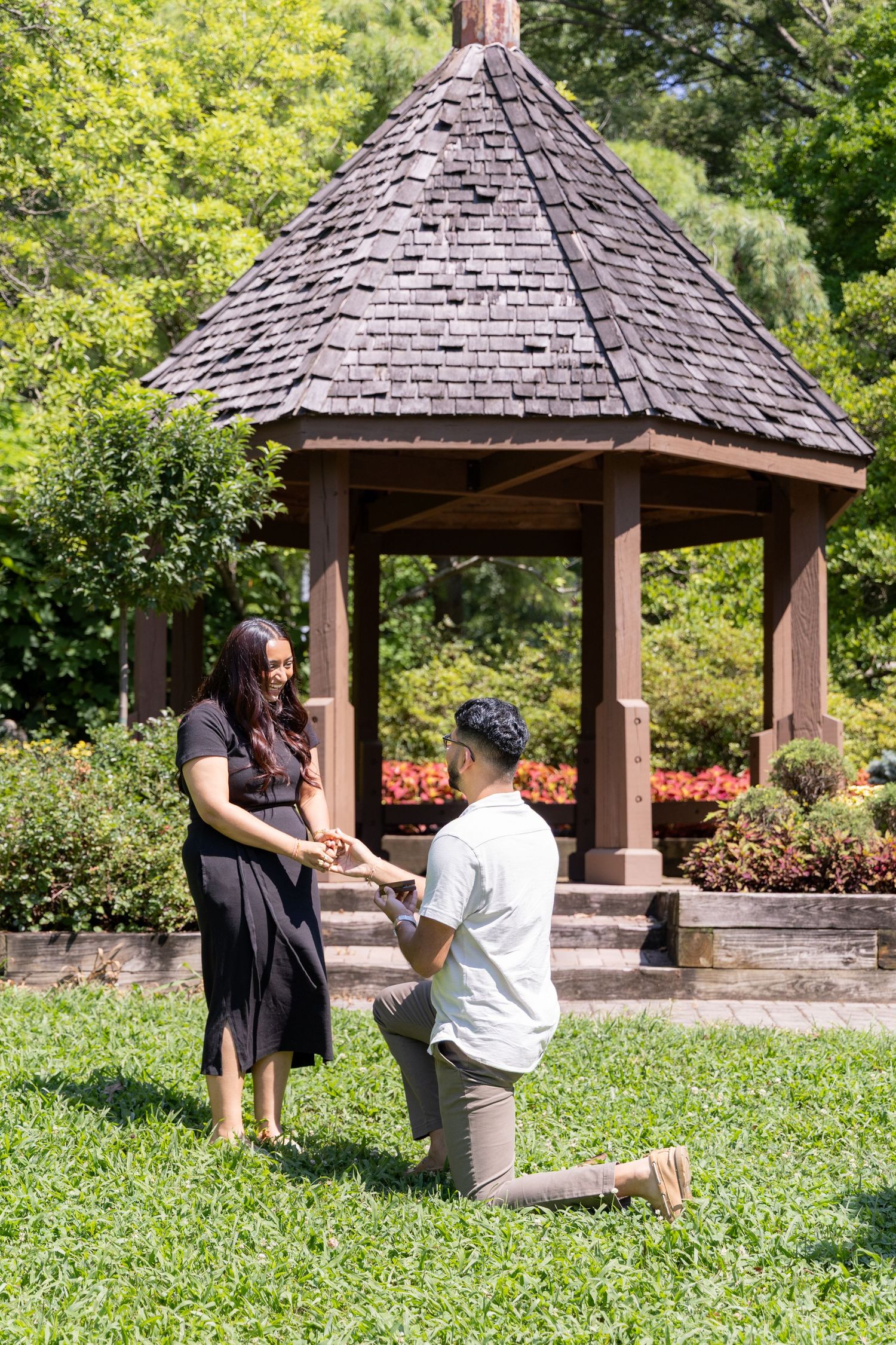 Man proposing on one knee to woman in front of a wooden gazebo surrounded by lush garden greenery at the Magnolia Garden in Eden Park.