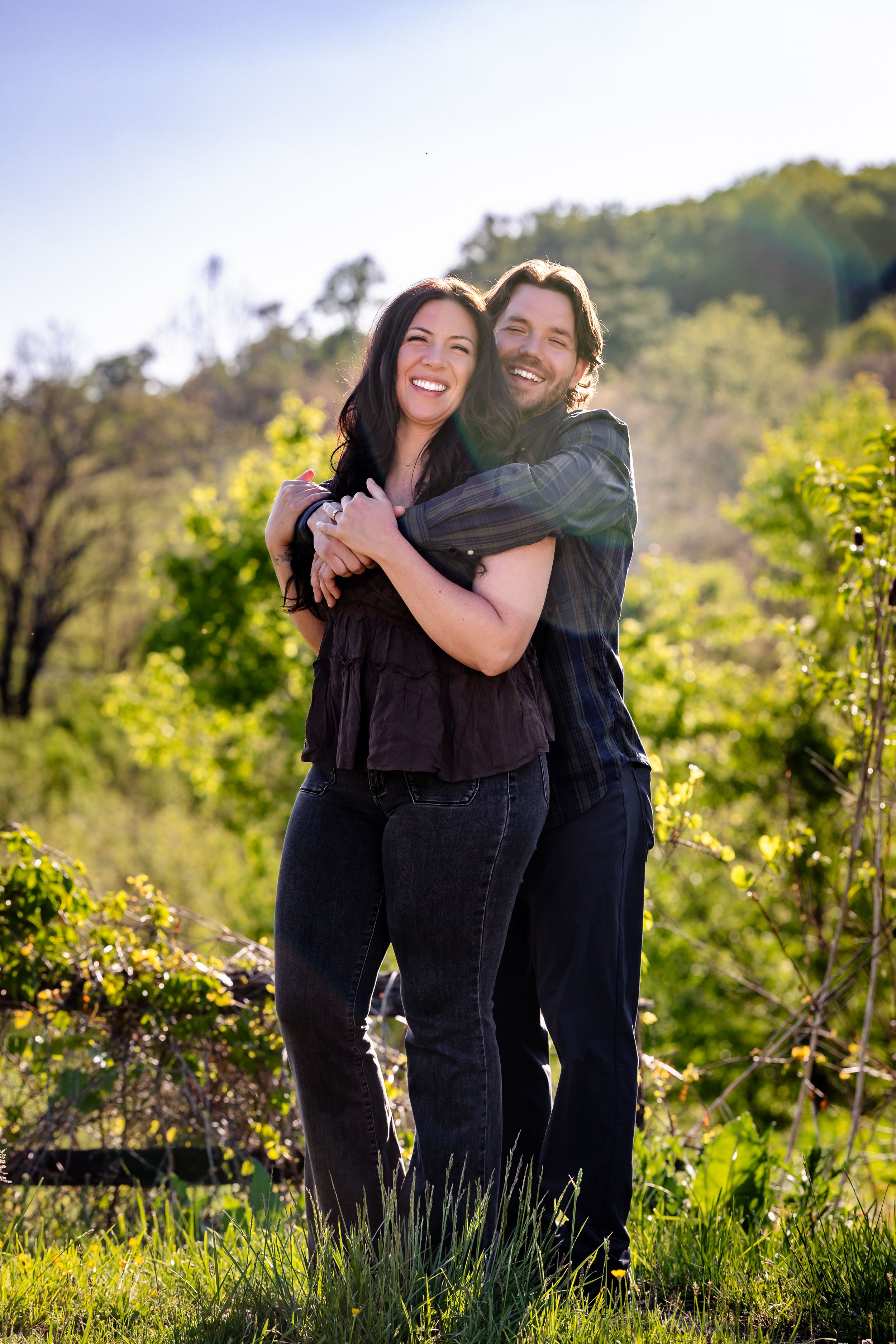 Happy couple embracing outdoors in a lush green garden, smiling in natural sunlight with trees in the background.