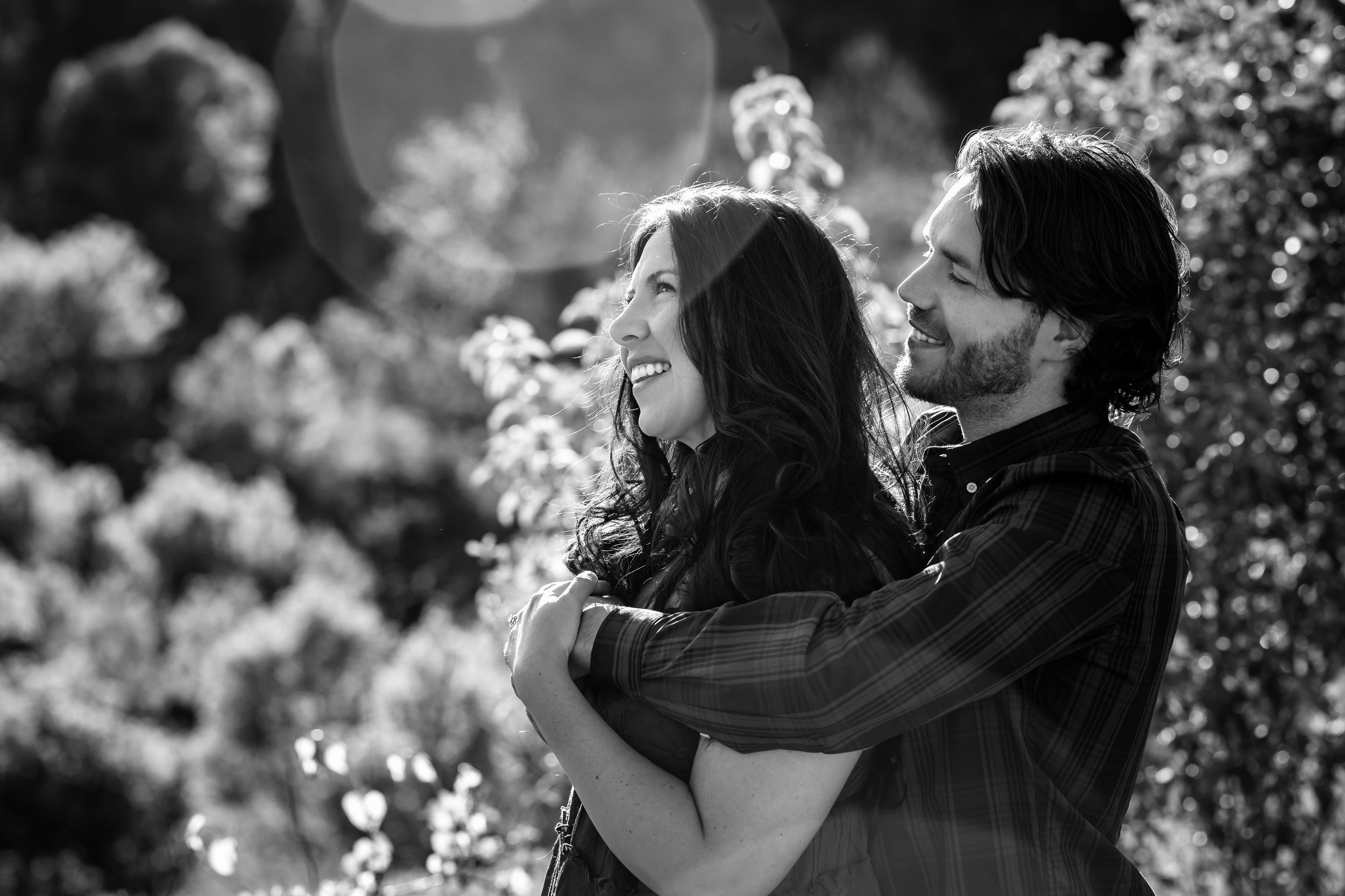 Black and white photo of a couple embracing and smiling outdoors among lush trees in a romantic moment.