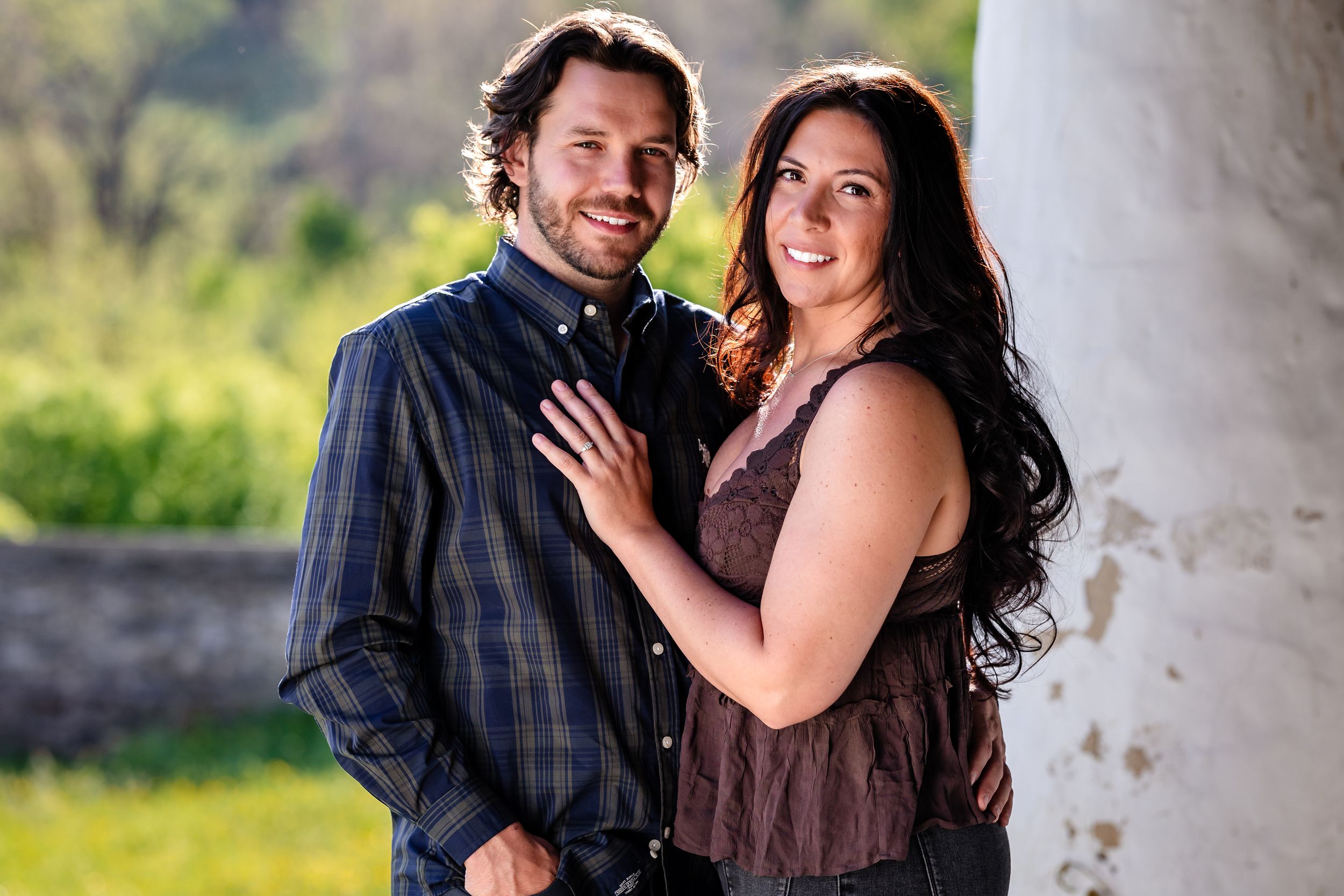 A smiling couple poses outdoors, man in plaid shirt embracing woman with long dark hair, green trees in background.