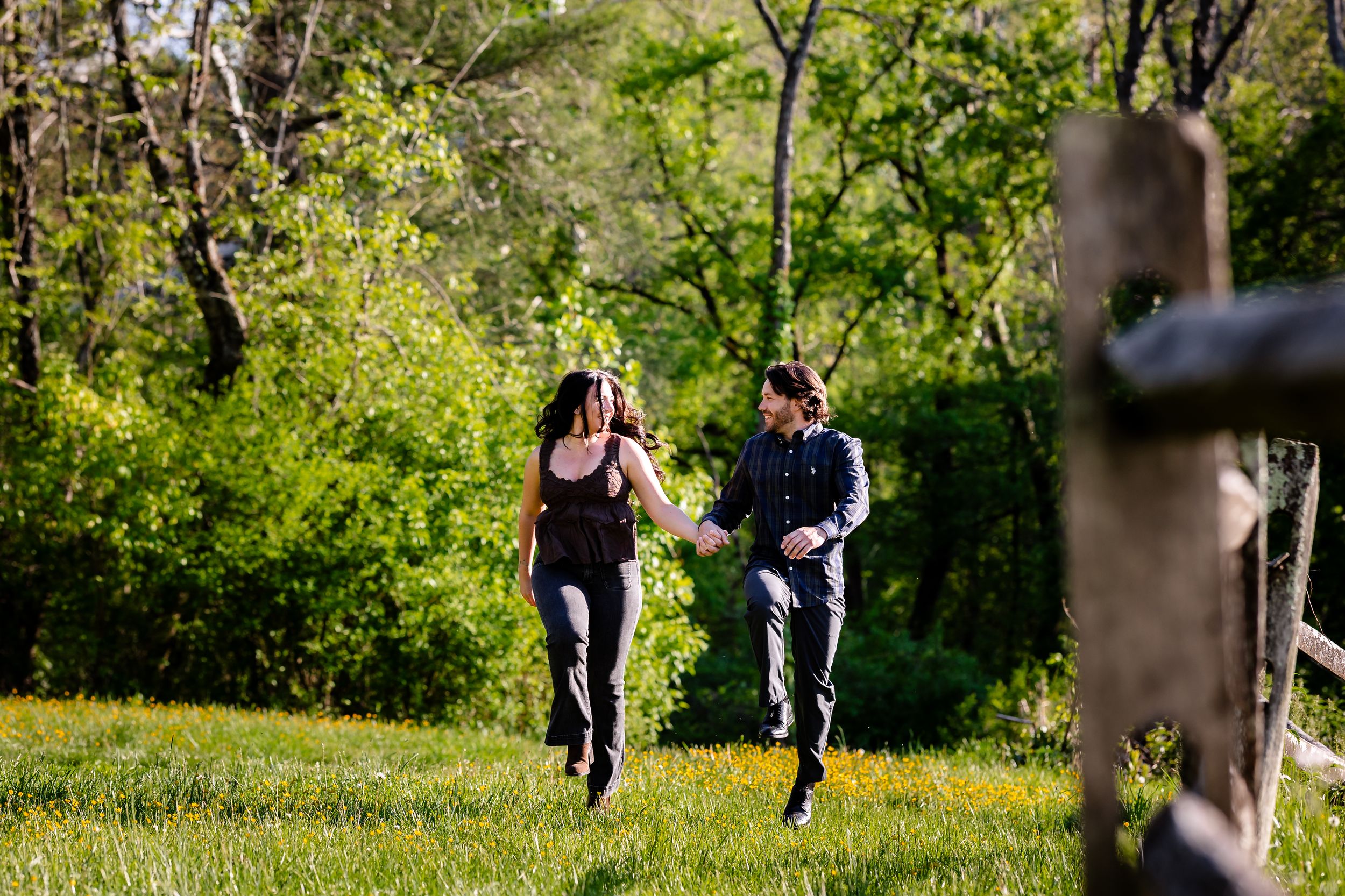 Couple holding hands and running through a lush green meadow surrounded by trees on a sunny day.