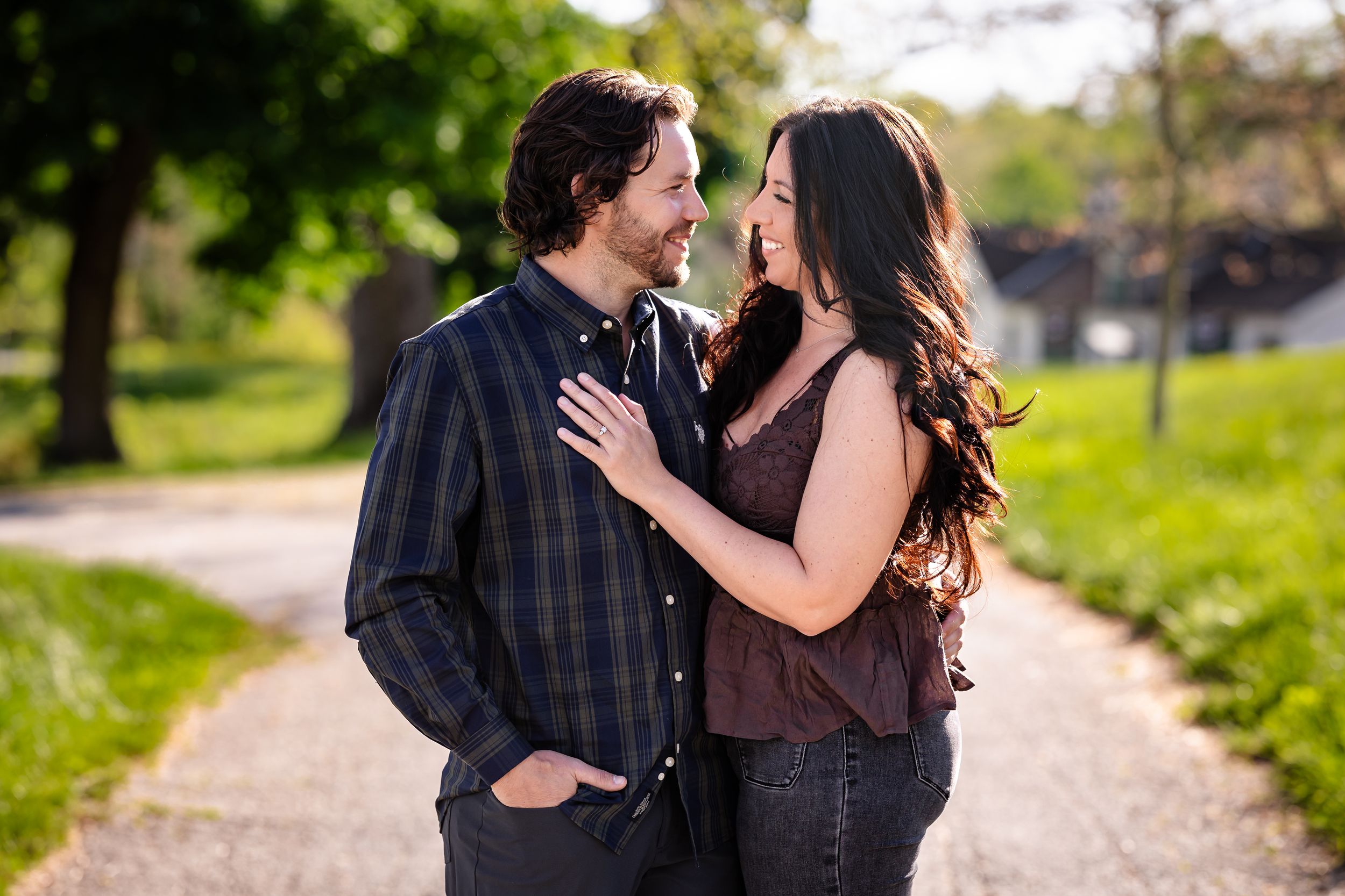 A couple shares a romantic moment on a sunlit path, smiling at each other in a lush green outdoor setting.
