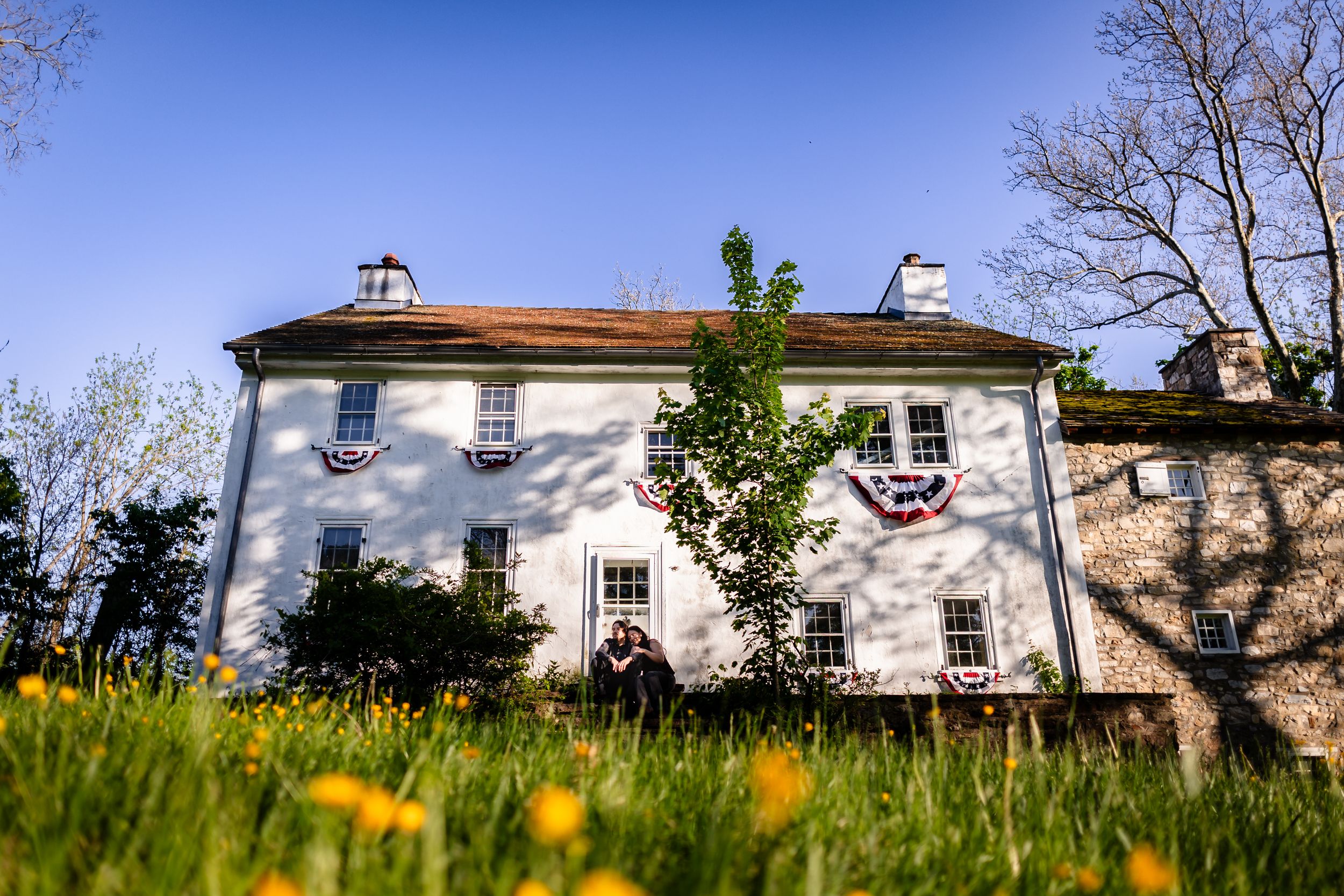 White colonial farmhouse decorated with patriotic bunting flags surrounded by wildflowers and trees under a clear blue sky.