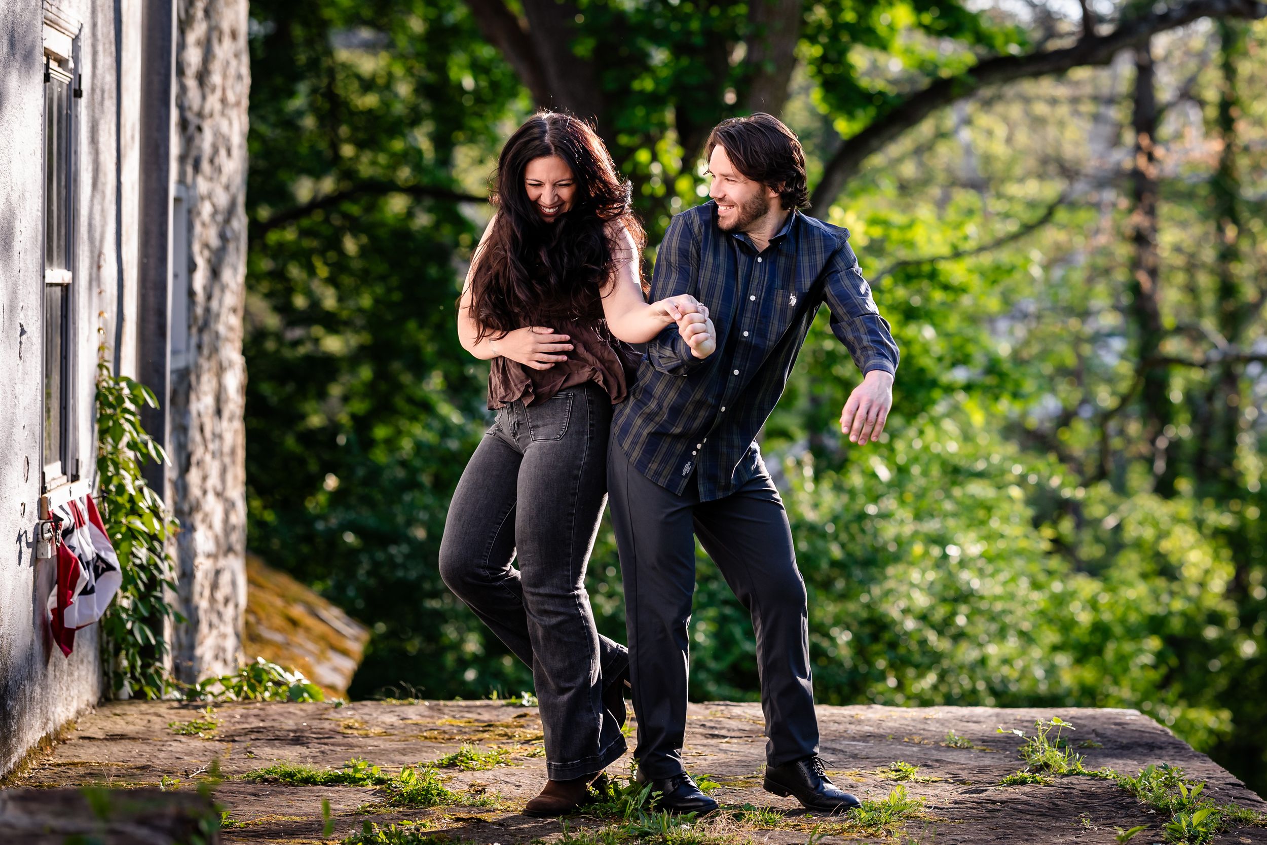 A couple laughing and dancing together outdoors, surrounded by lush green trees in a natural setting.