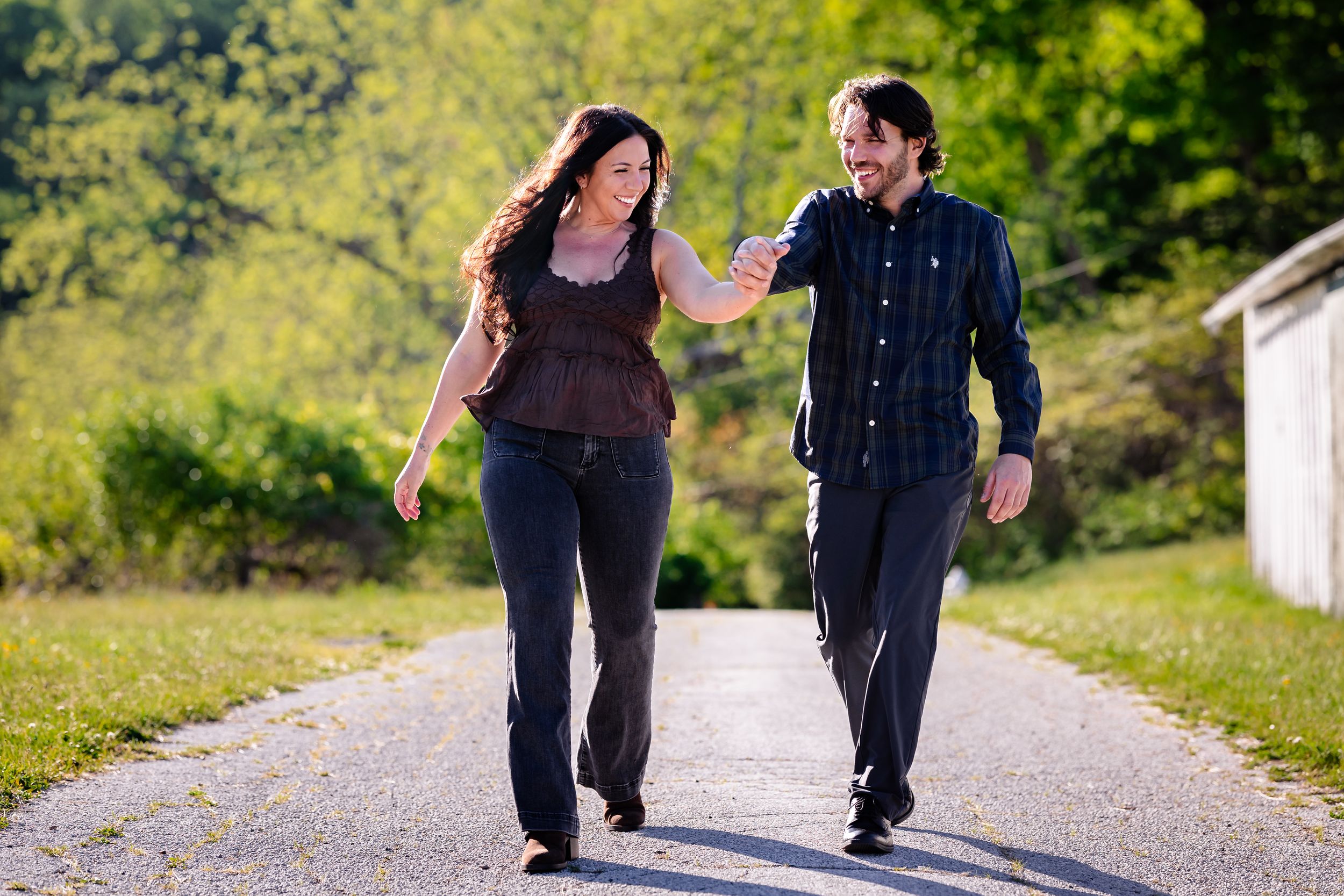 Couple walking hand-in-hand on a sunny path surrounded by lush green trees, smiling and enjoying a romantic outdoor stroll.