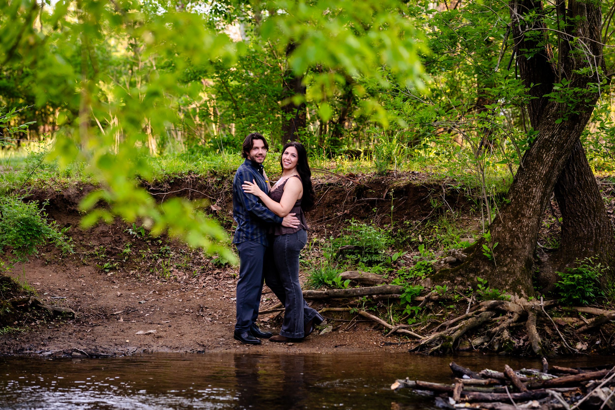 Couple embracing romantically by a serene forest creek surrounded by lush green trees during an outdoor engagement session.