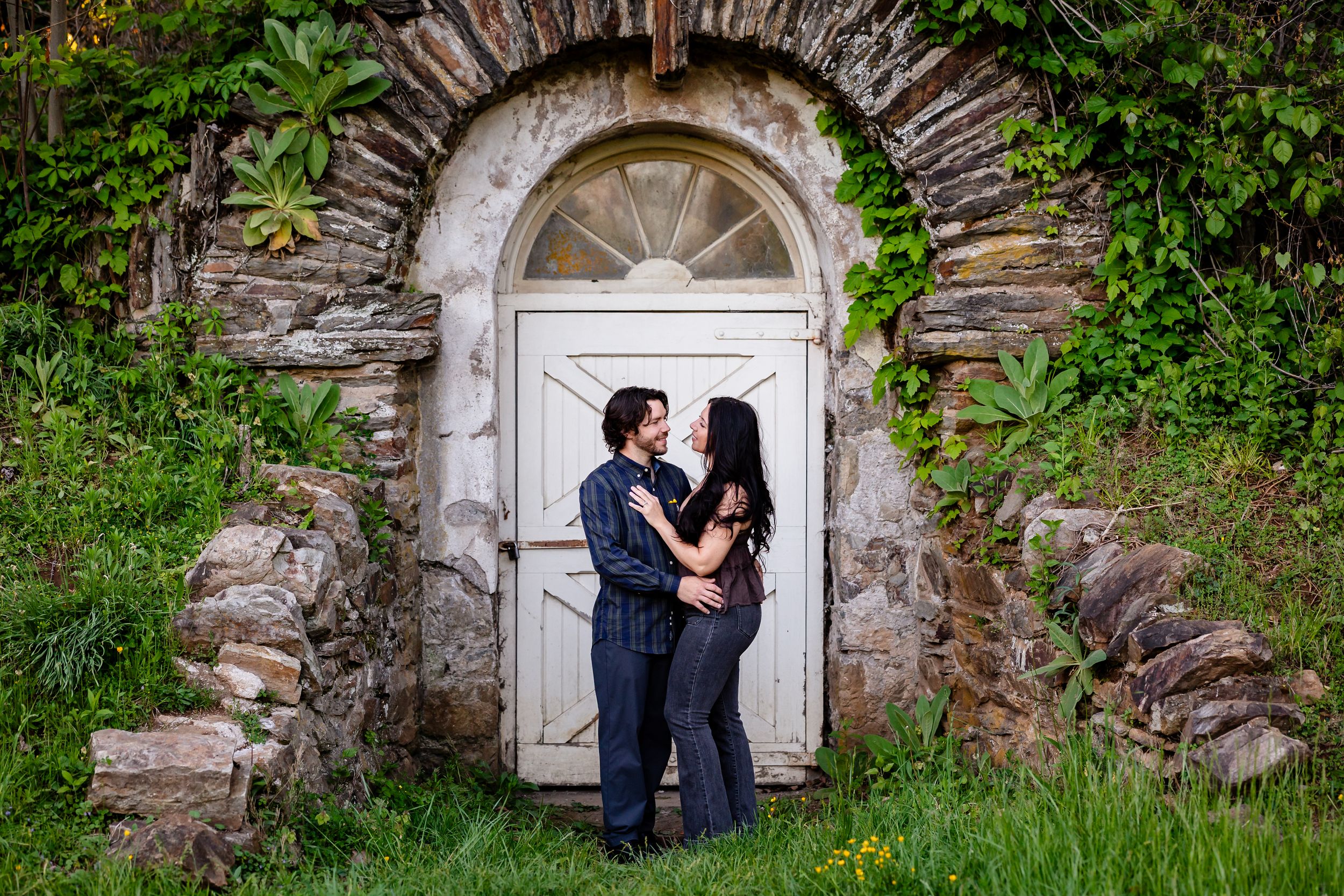 Couple embracing in front of a rustic stone archway with a white door, surrounded by lush greenery.