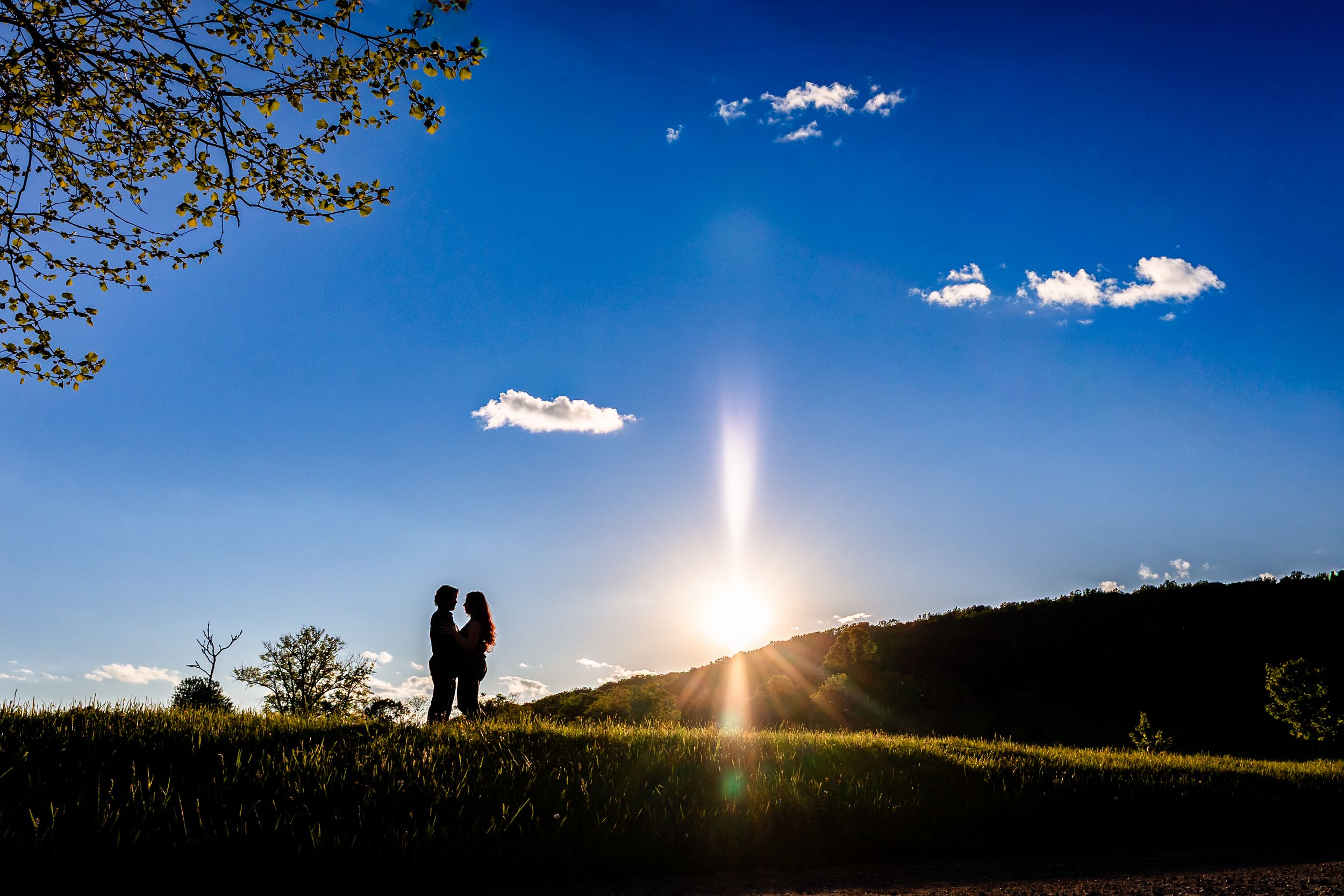 Romantic couple silhouette at golden sunset with sun flare on a green hillside under a vivid blue sky.