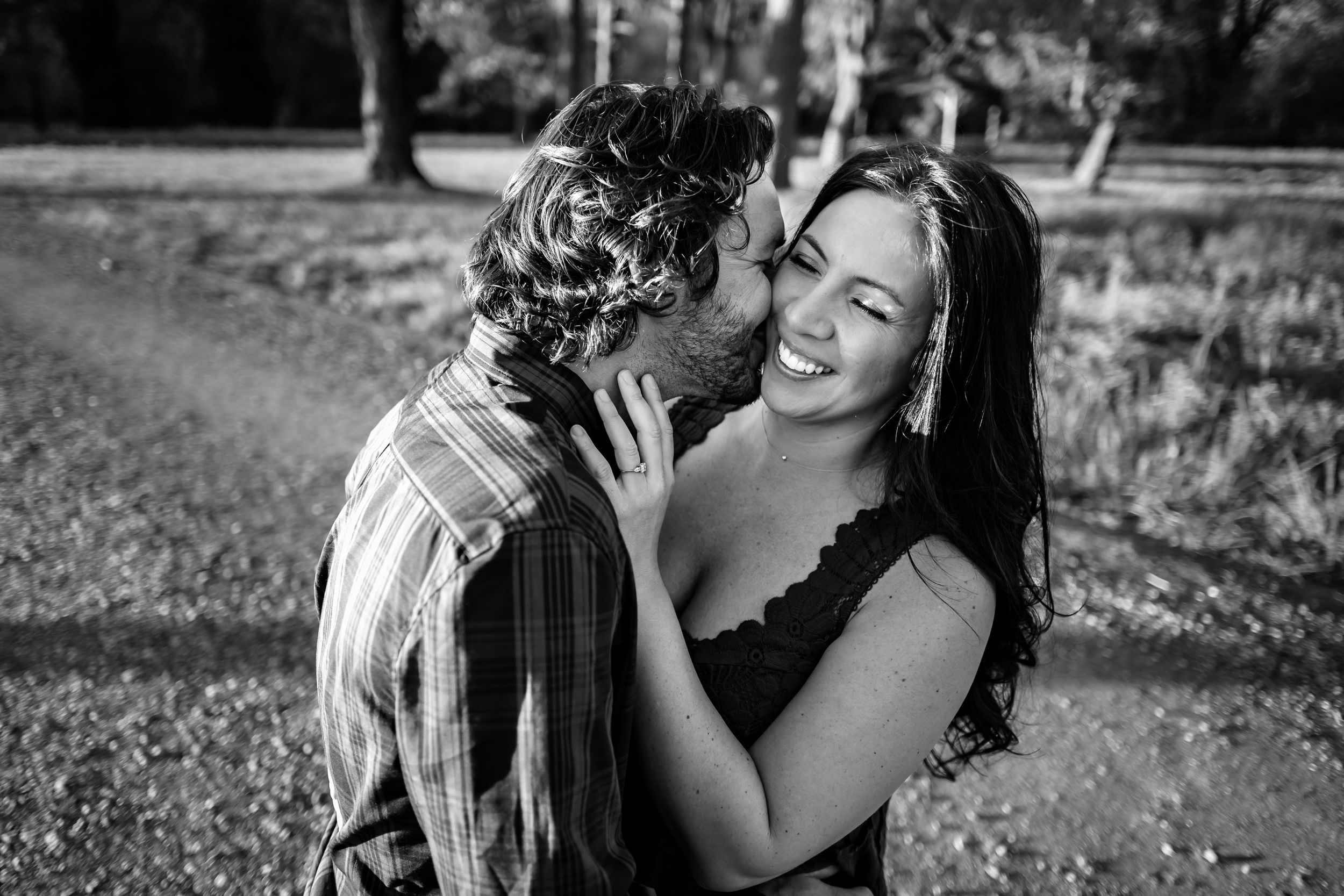 Black and white photo of a couple sharing a romantic moment outdoors, woman smiling joyfully as man kisses her cheek.