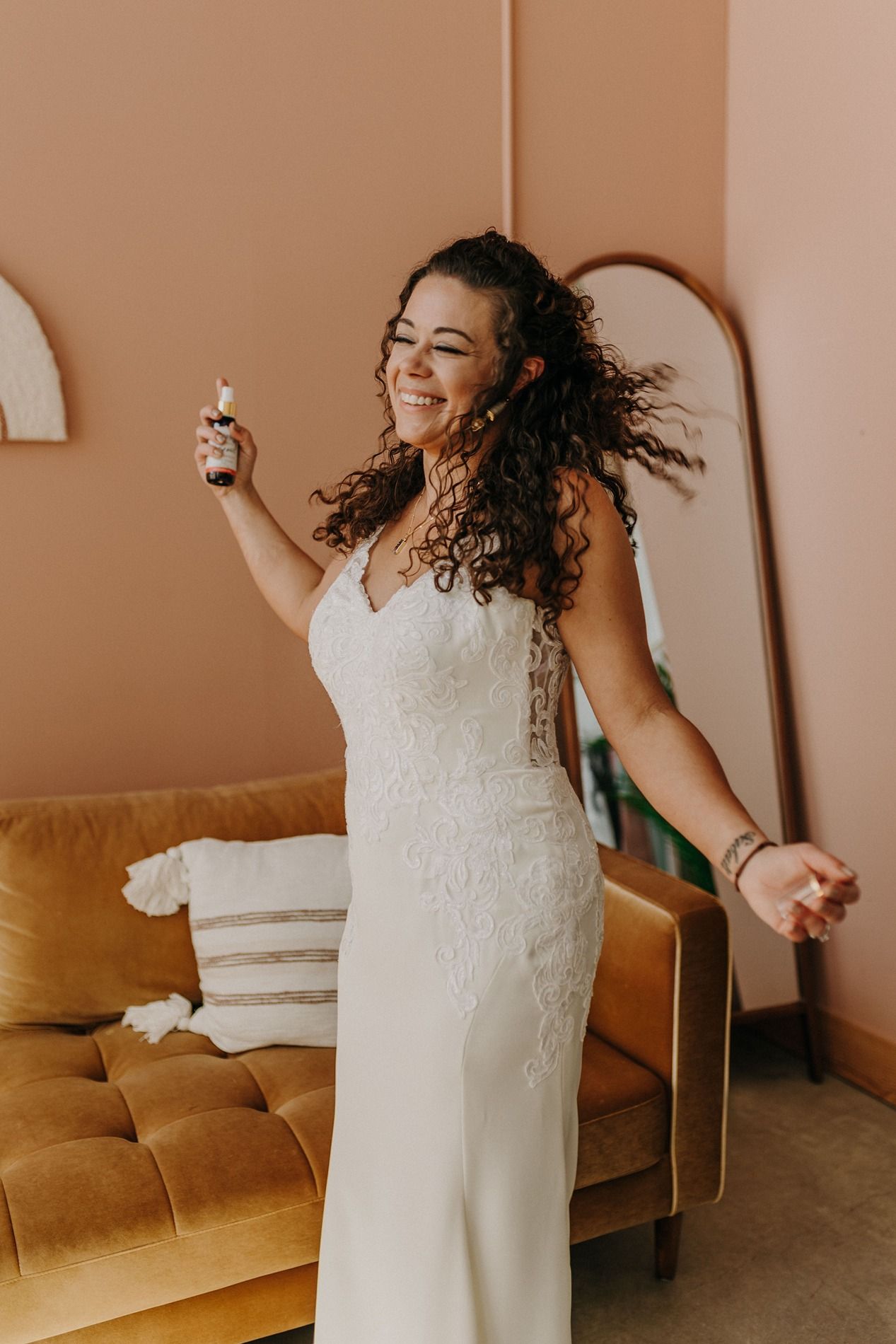 Joyful bride in white lace wedding gown dancing and smiling, holding a bottle, with curly hair flowing at The Lane.