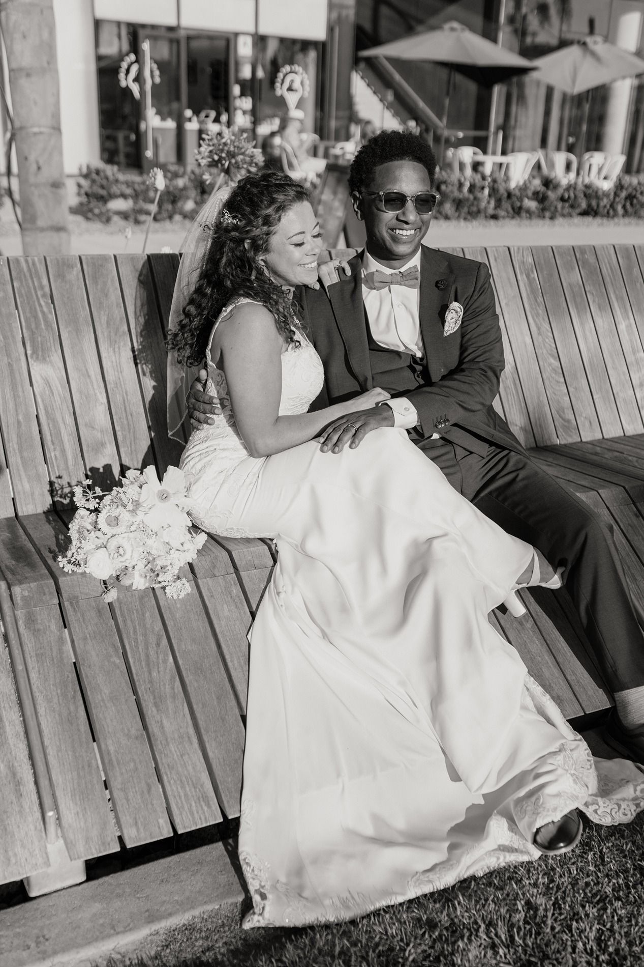 Black and white photo of a bride and groom sitting on a wooden bench, smiling, with a bouquet of flowers at The Lane.