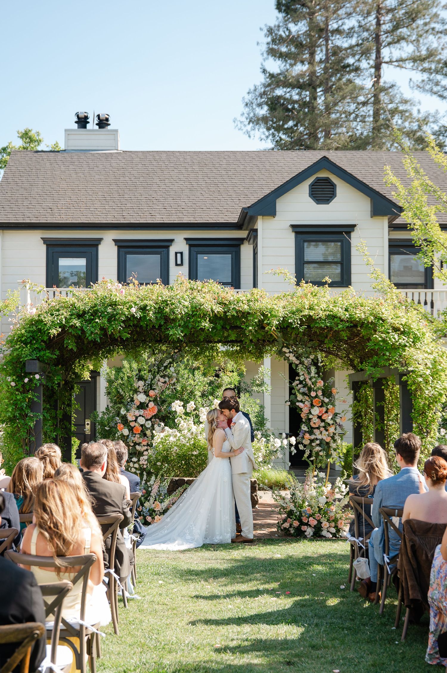 Outdoor wedding ceremony with couple kissing under floral arch, guests seated on lawn before elegant vine-covered white home.