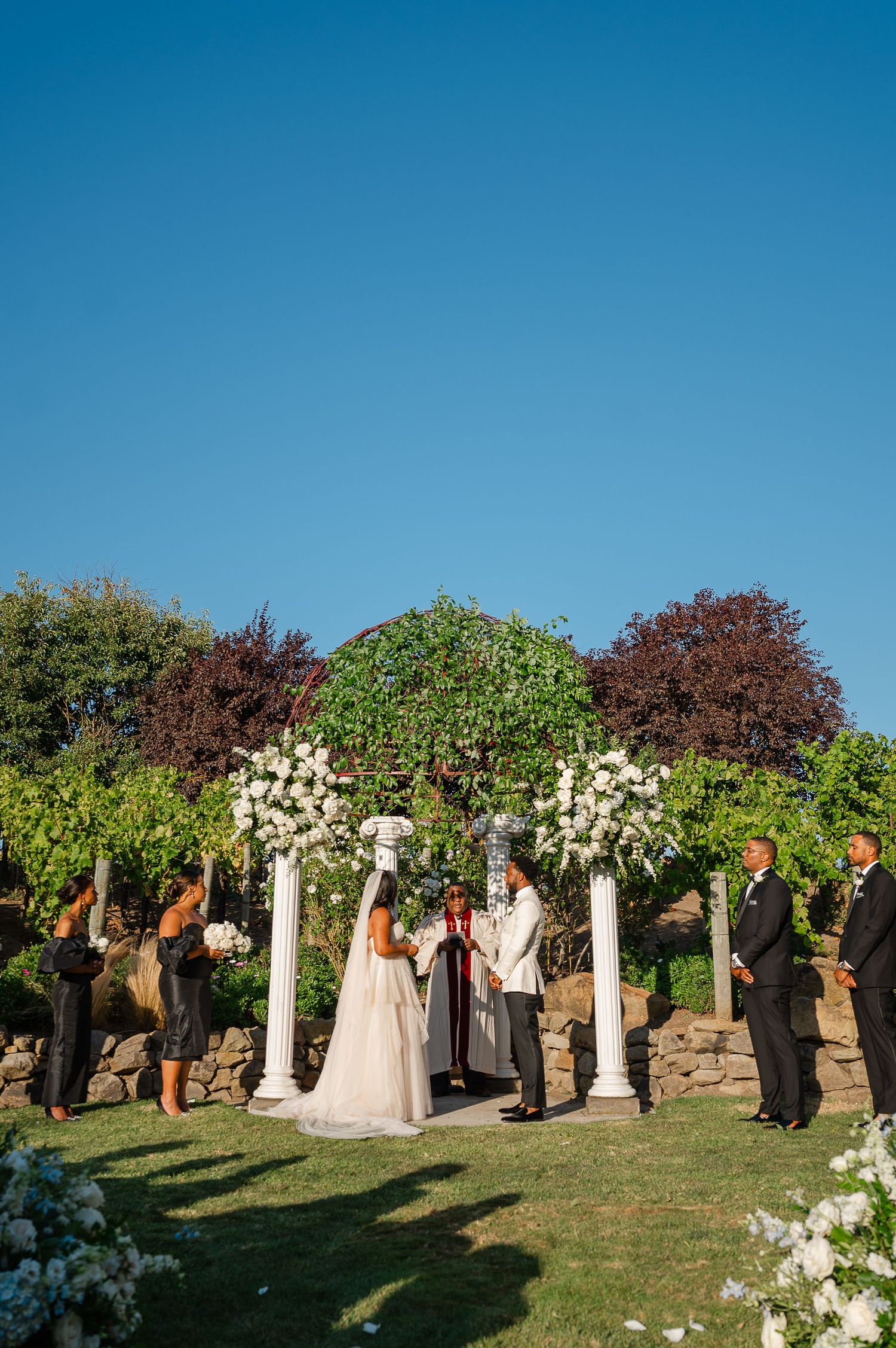 Bride and groom exchange vows under a white floral arch during an outdoor wedding ceremony.