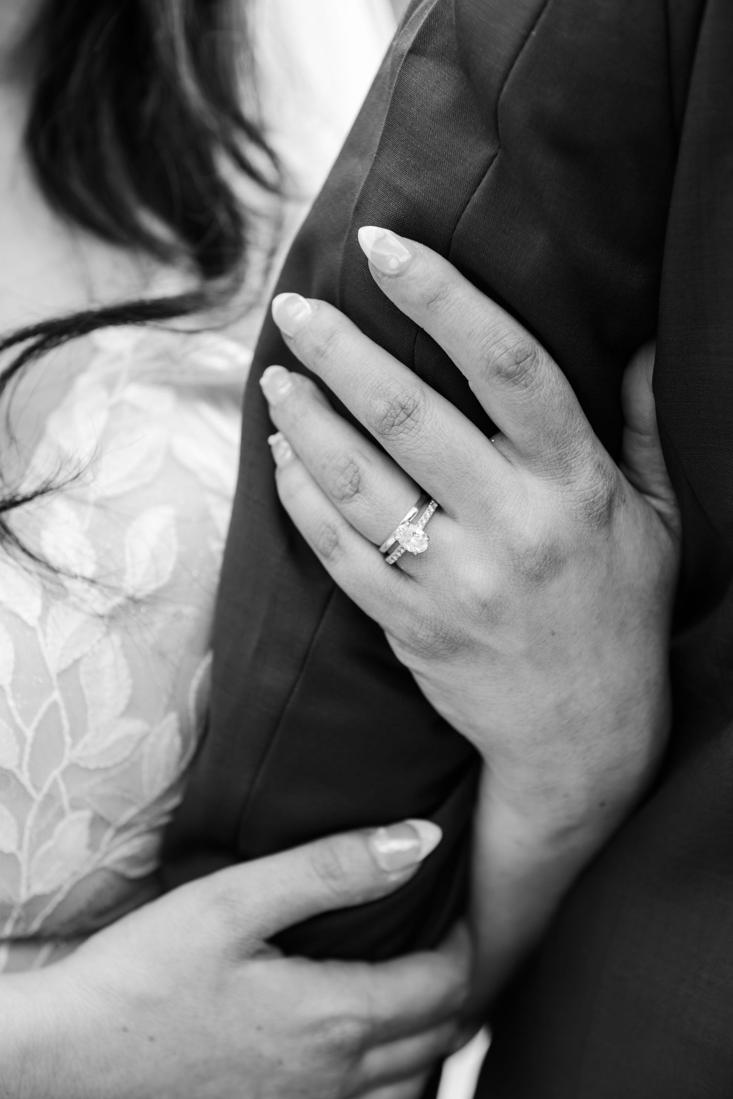 Black and white photo of a bride's hand with diamond engagement ring resting on groom's suit jacket.