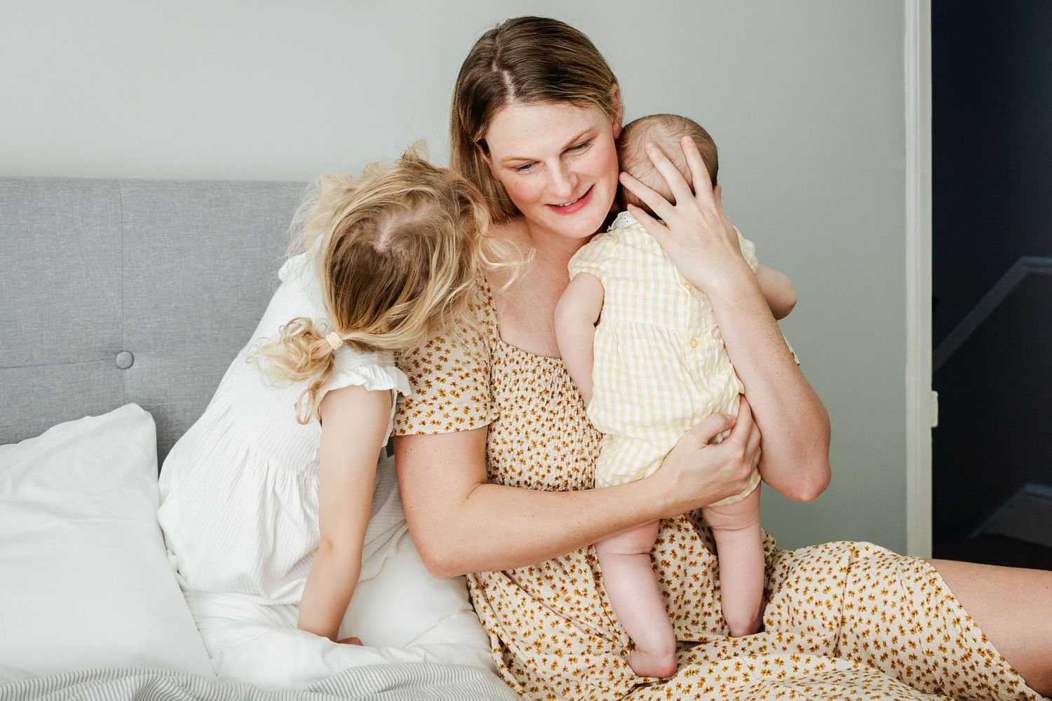 A mother sits on her bed at home and holds her baby to her chest. Her older daughter whispers in her ear. Bristol family photographer