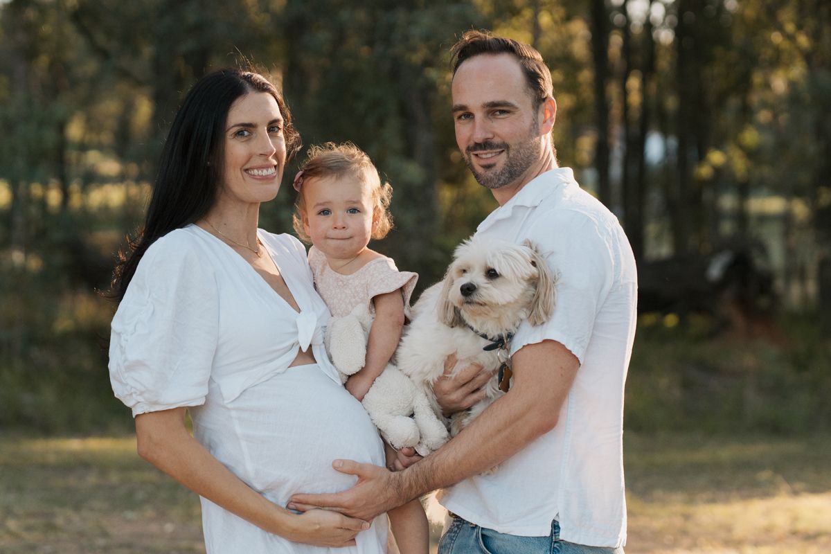 Pregnant woman and man in white outfits holding toddler and small white dog outdoors in a park in Rouse hill family photoshoot with a dog
