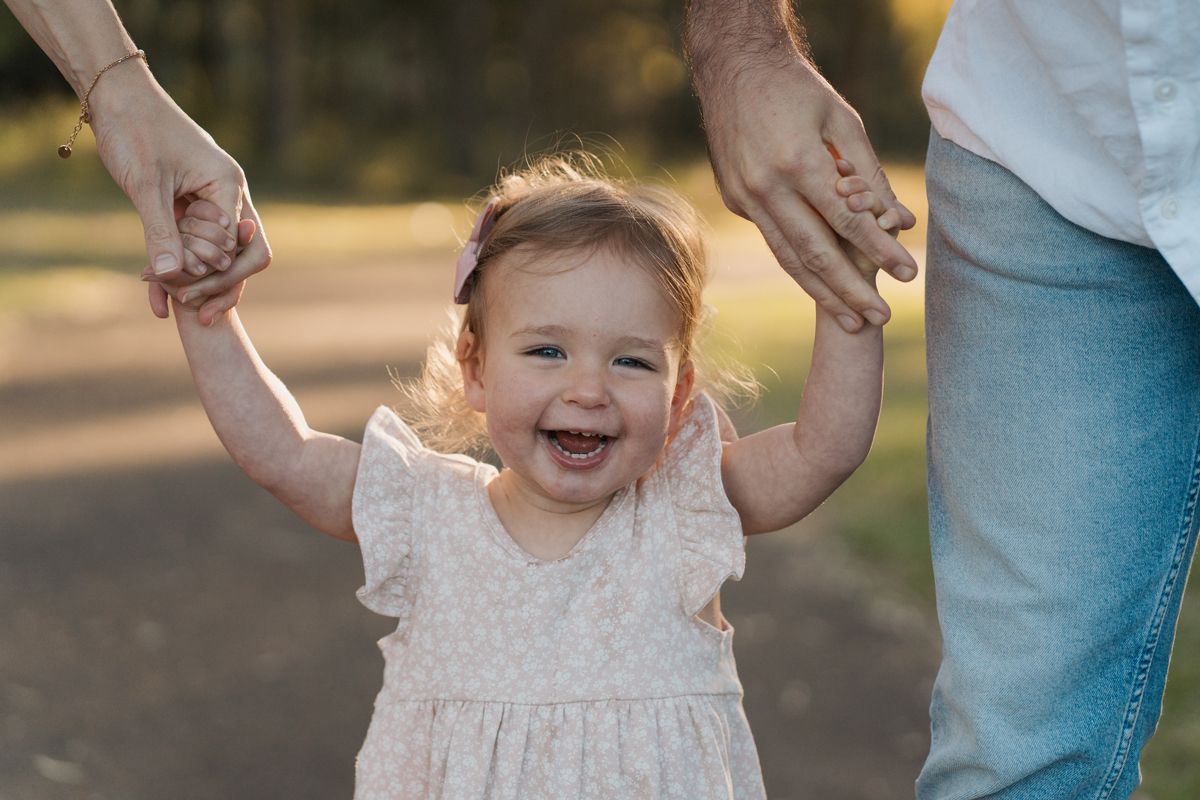Smiling toddler girl in white dress joyfully holds hands with parents while walking outdoors in warm sunlight in rouse hill family photoshoo