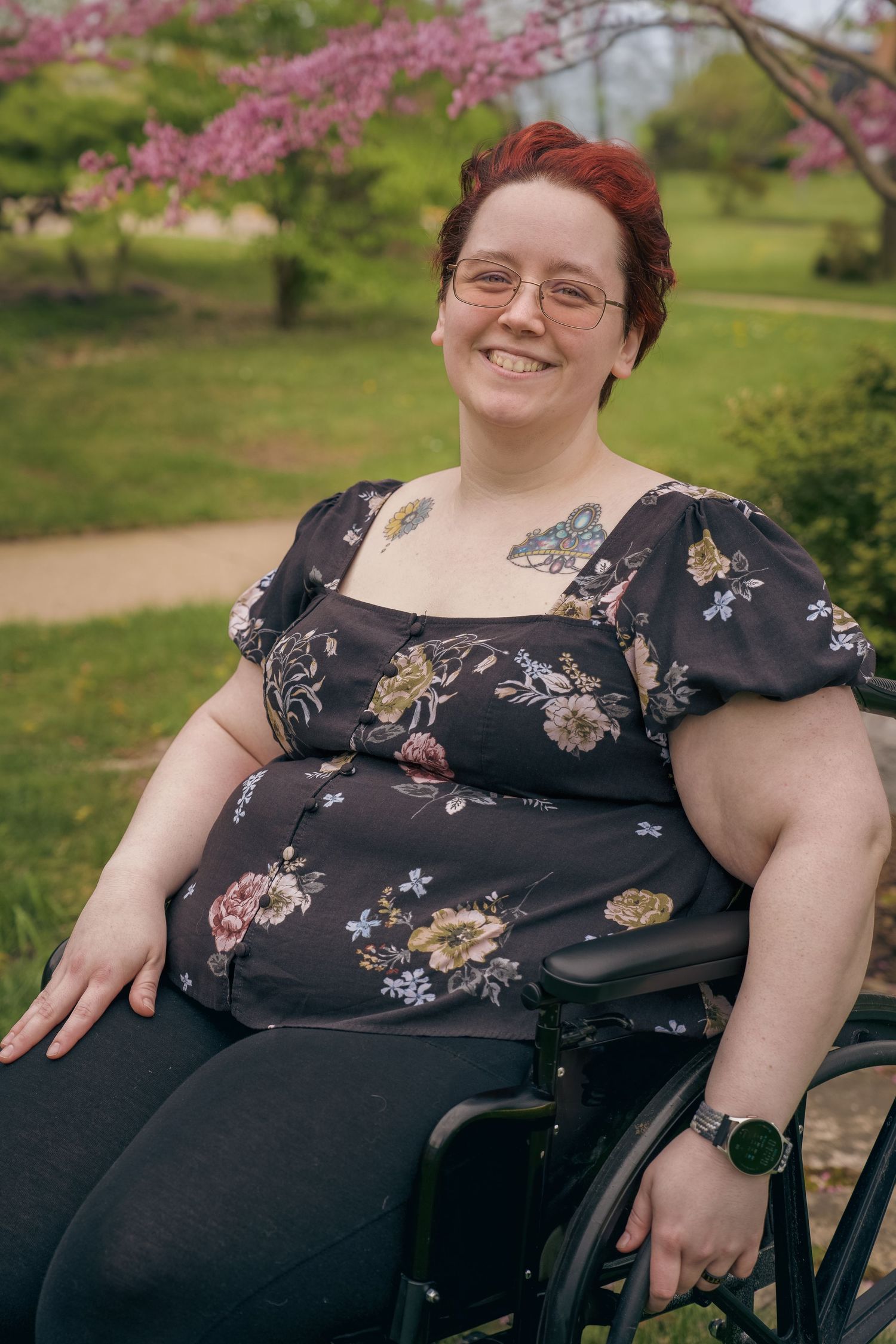 Smiling woman with red hair and glasses sitting in a wheelchair outdoors near a blooming pink tree.