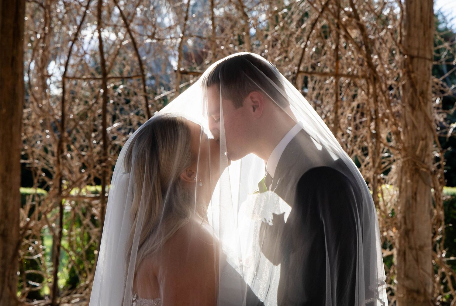 Bride and groom share a kiss at an elegant outdoor pavilion wedding venue with wooden cross-back chairs.