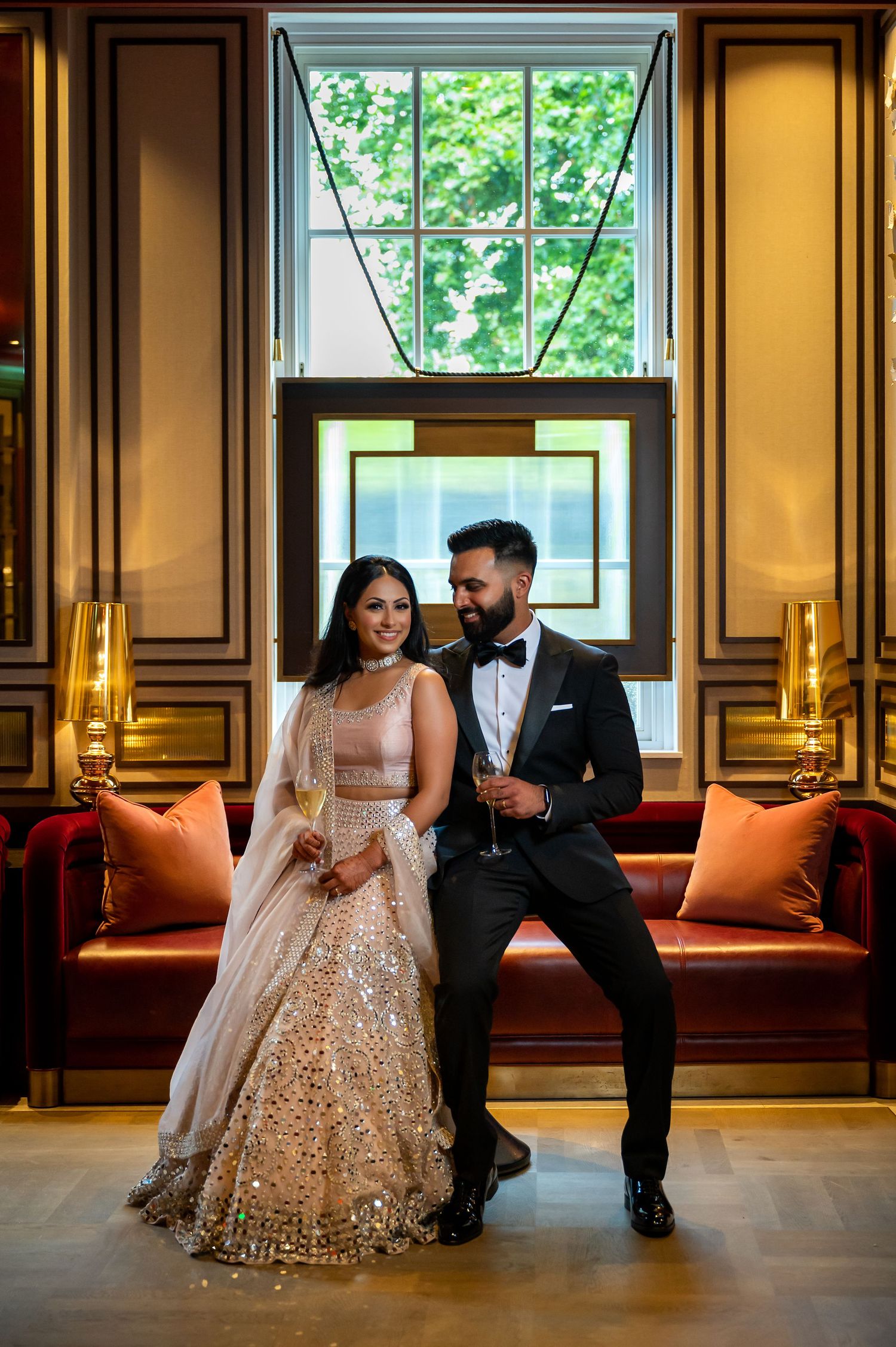 Couple in elegant wedding attire holding champagne flutes, woman in embellished lehenga, man in black tuxedo, luxury hotel interior.