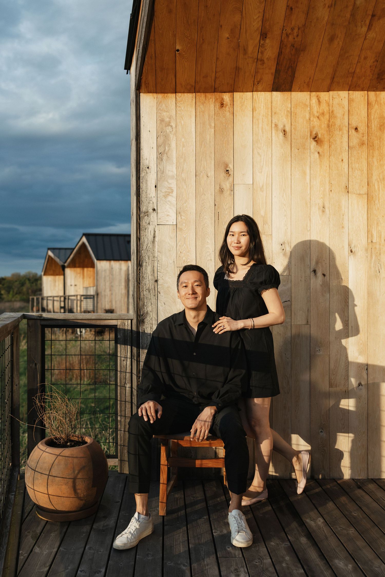 A couple poses on a wooden deck balcony, man seated on stool, woman standing beside him, against a rustic wood wall.