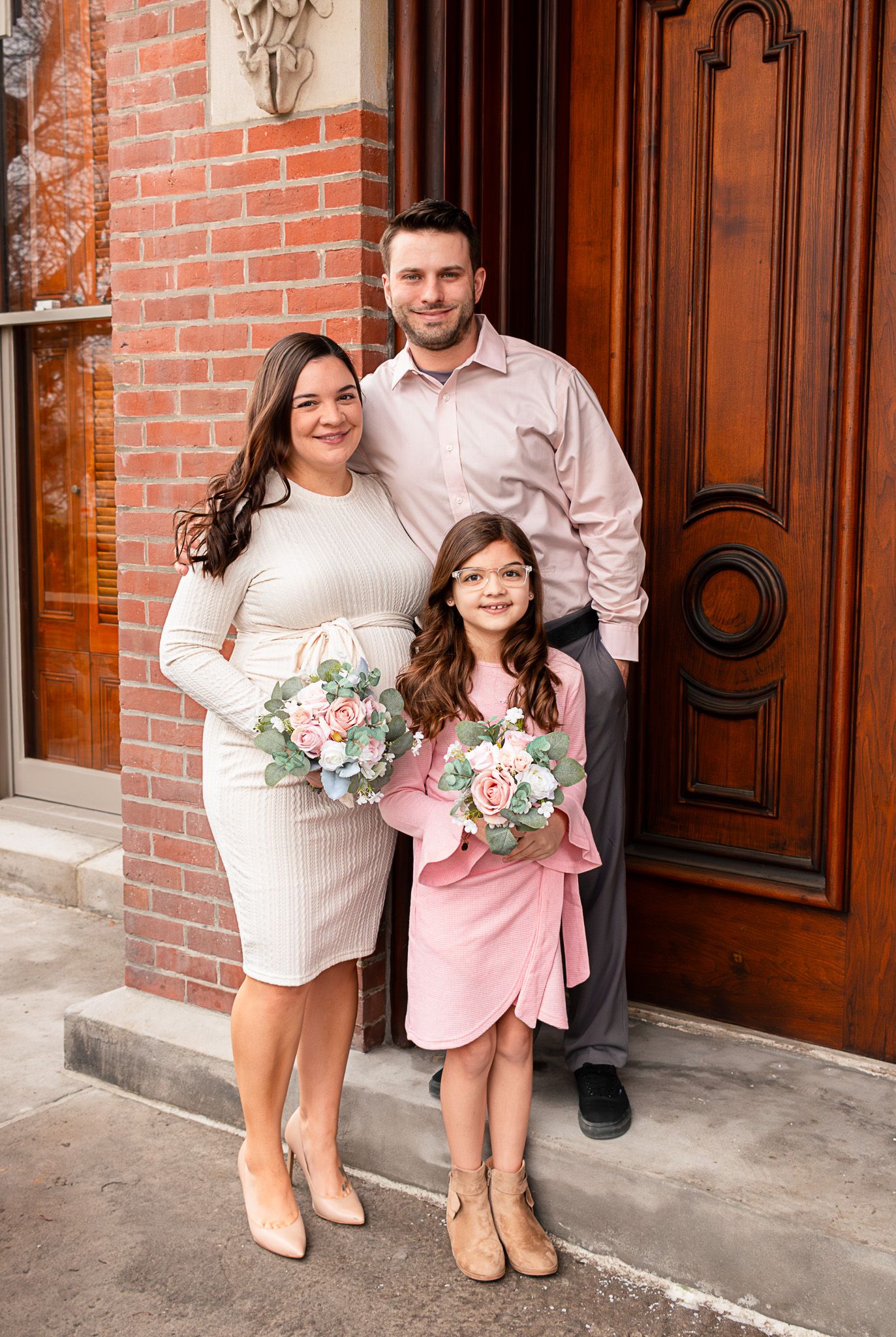 Family of three posing by ornate wooden door, woman and girl holding floral bouquets, dressed in elegant attire.
