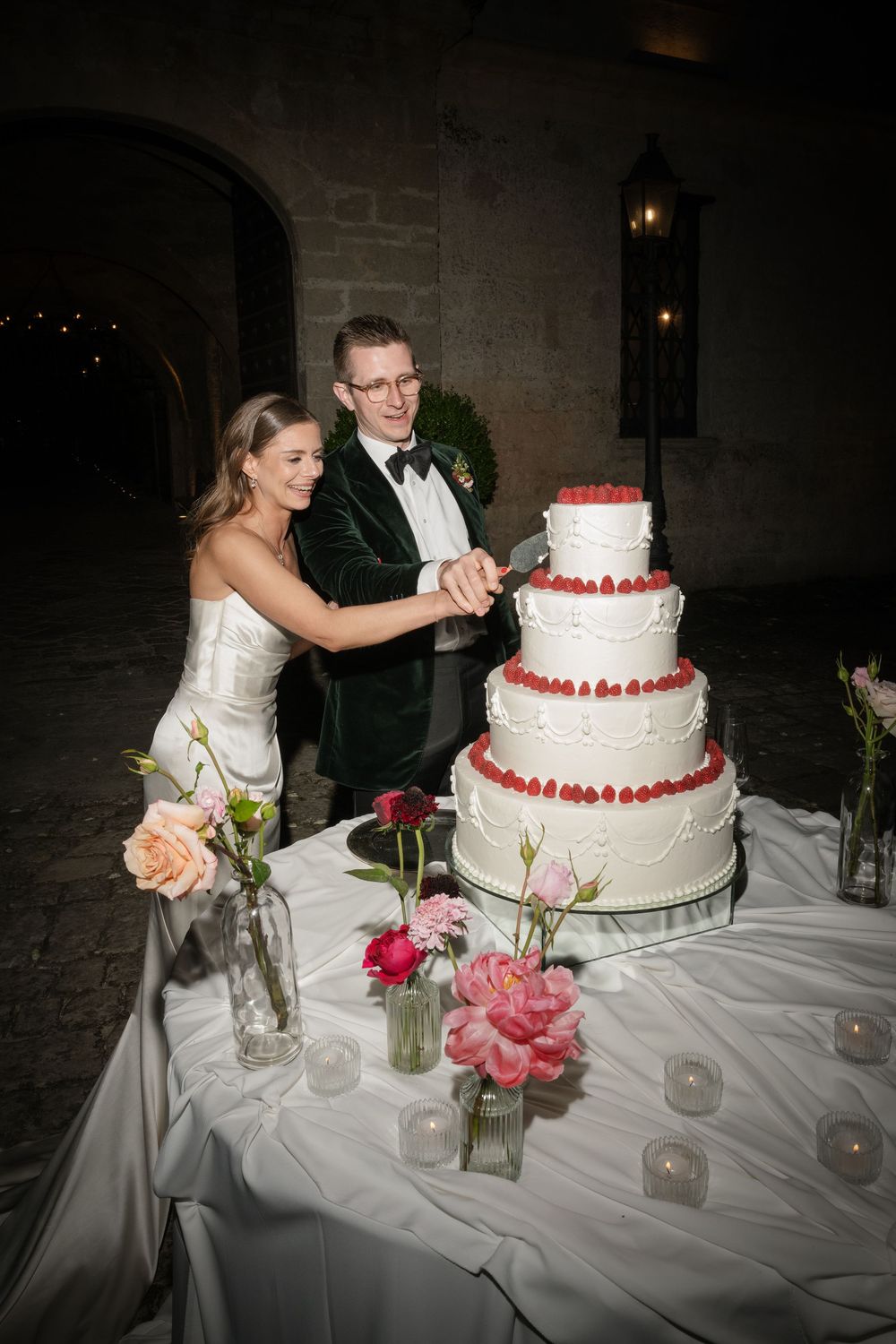 Bride and groom cutting a four-tier white wedding cake decorated with raspberries, surrounded by floral arrangements and candles.