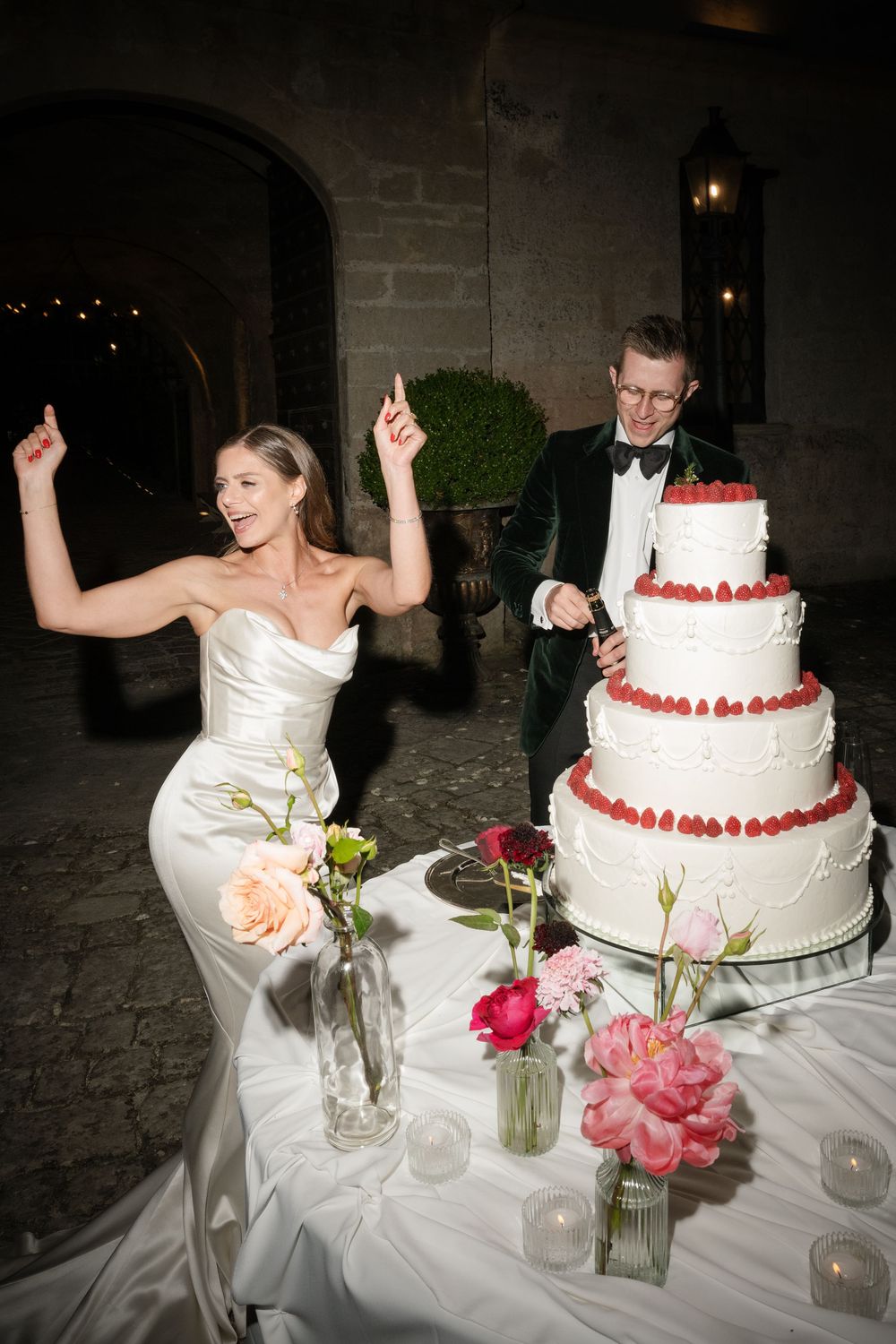 Bride in white strapless gown celebrates with arms raised beside groom cutting four-tier wedding cake with strawberries.