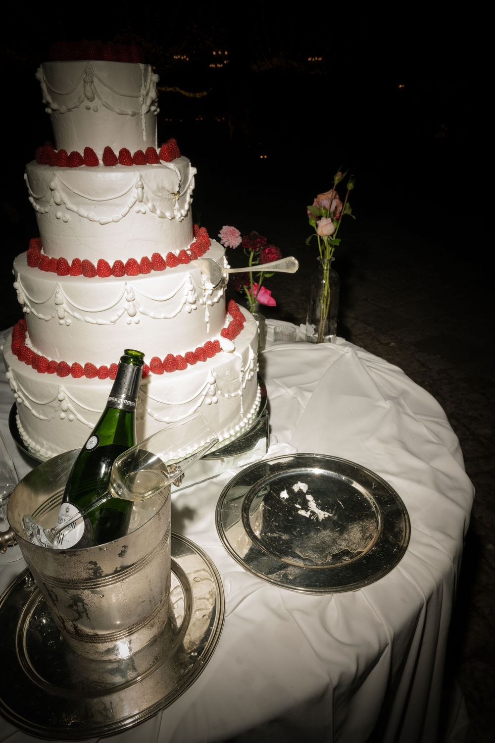 Four-tier white wedding cake with strawberry decorations beside champagne on ice at night.