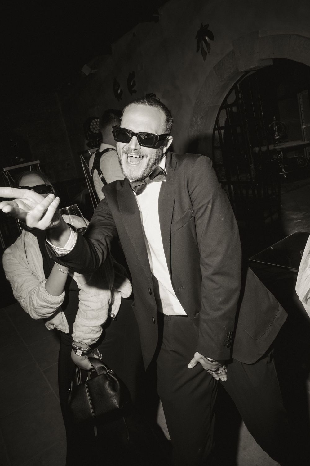 Man in tuxedo and sunglasses smiling at a formal event in black and white photo.