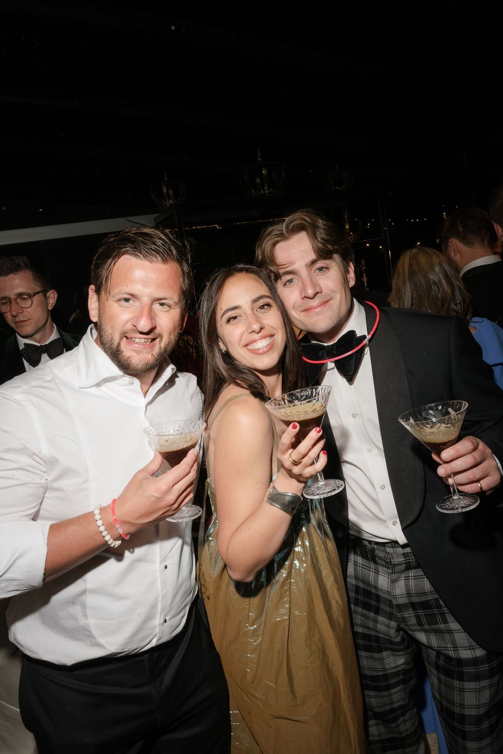 Three friends smiling and holding cocktails at a formal evening party event.