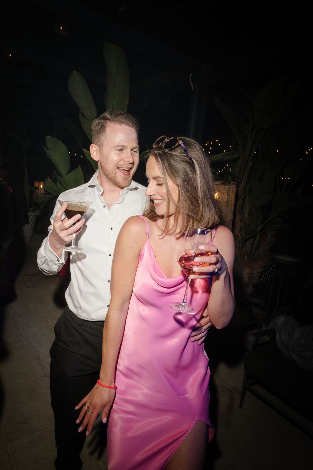 A couple in elegant attire enjoying cocktails at an outdoor evening party, woman in pink satin dress smiling.