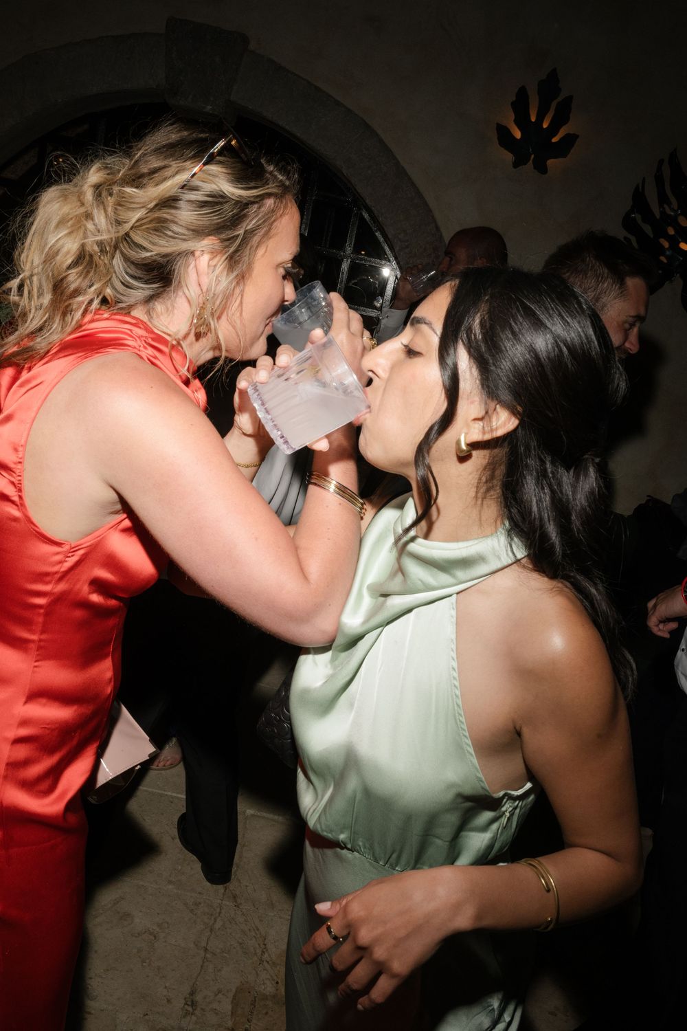 Two women in elegant dresses share a drink at a glamorous party event.