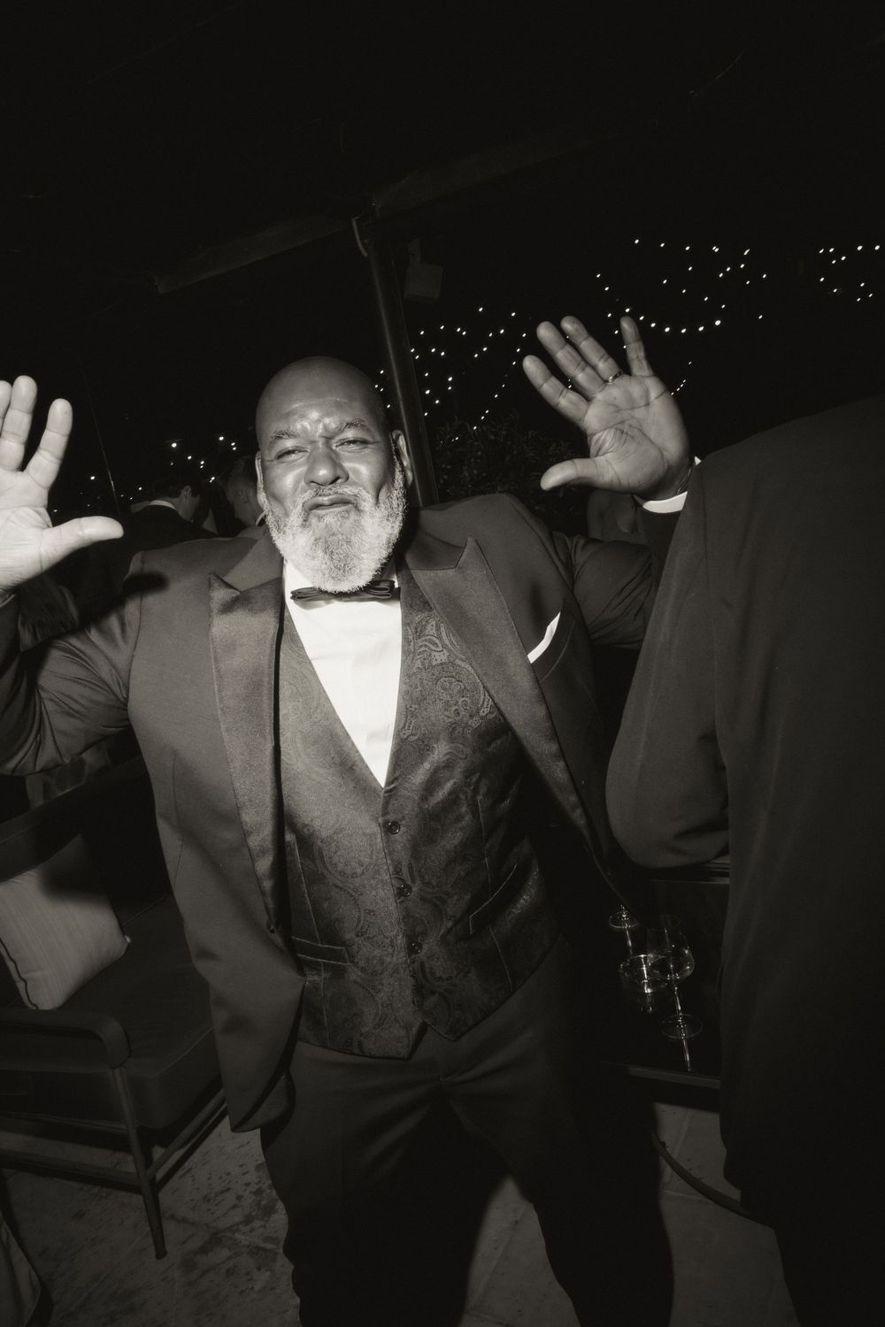 Elderly man in tuxedo with bow tie dancing with hands raised at formal evening event.