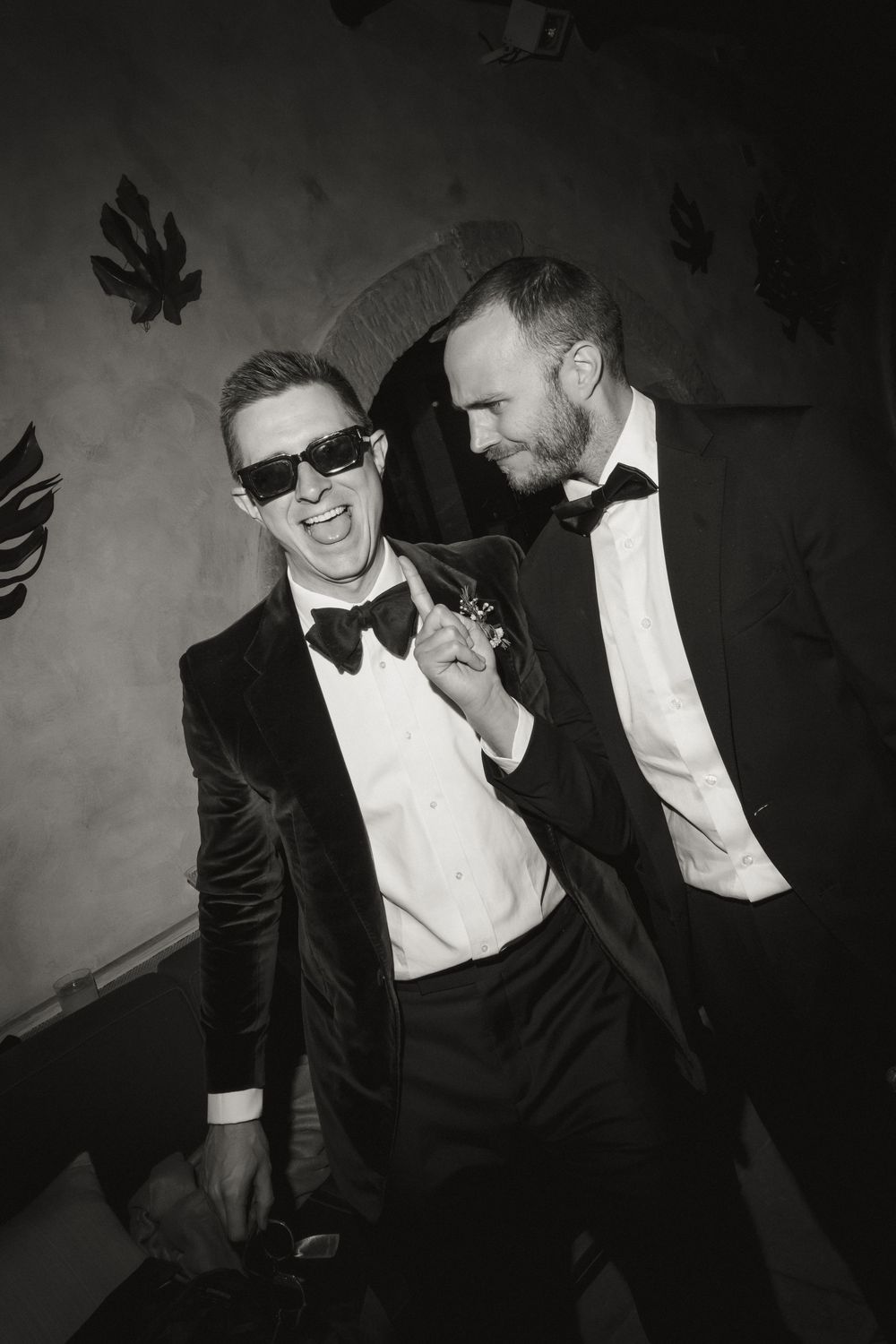 Two men in tuxedos and bow ties laugh and pose together in a black and white formal event photo.