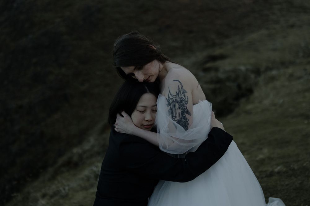 Two people sharing a tender embrace outdoors on a misty hillside, one wearing a white wedding dress with a floral tattoo.
