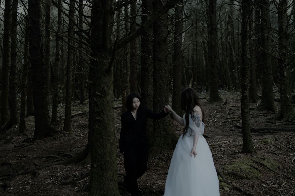 Two figures in dark forest, one in black mask and suit, one in white dress, creating eerie gothic atmosphere.