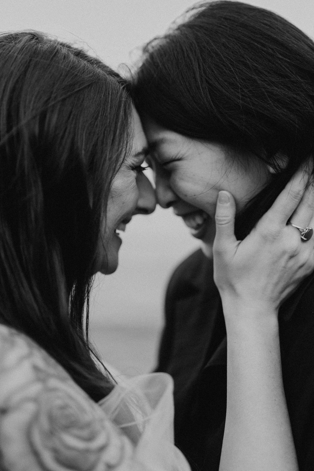 Black and white photo of two women laughing and touching foreheads, one wearing an engagement ring.