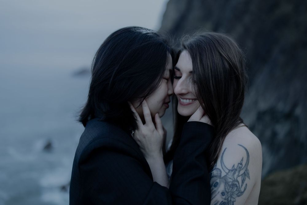 Two women with dark hair share an intimate moment outdoors near coastal cliffs, one with a tattoo on her shoulder.