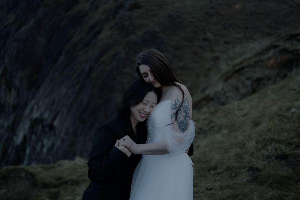 Two brides embrace tenderly on a dramatic rocky hillside, one in a white gown and the other in black.