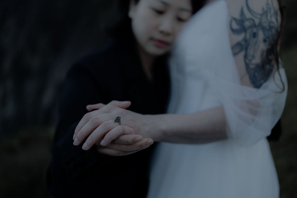 Two women holding hands, one in black and one in white dress with a floral tattoo, in an intimate outdoor setting.
