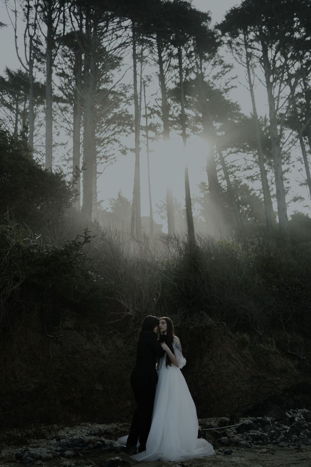 Couple embracing in misty forest at dusk, bride in white gown and groom in black suit, with sunbeams through tall trees.