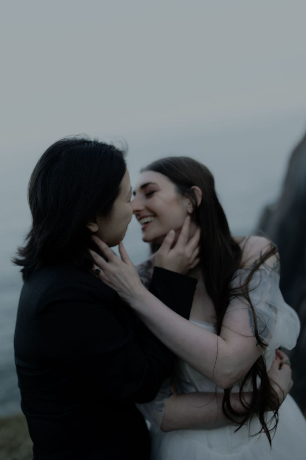Two brides share a tender almost-kiss outdoors, one in a black suit and one in a white dress, against a misty coastal backdrop.