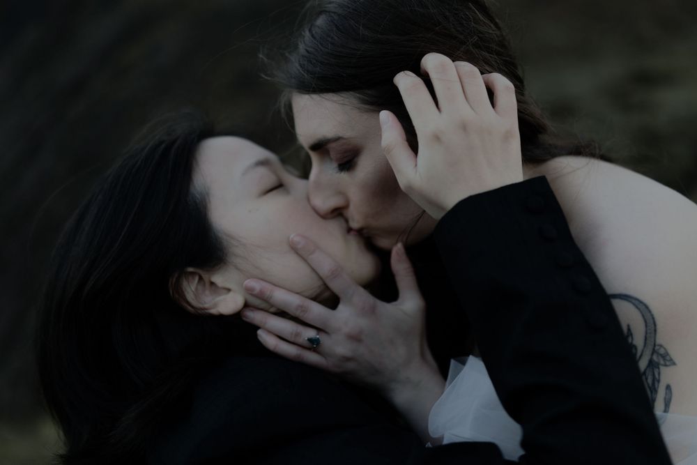 Two women sharing a romantic kiss, one with a tattoo on her arm, in a dark moody outdoor setting.