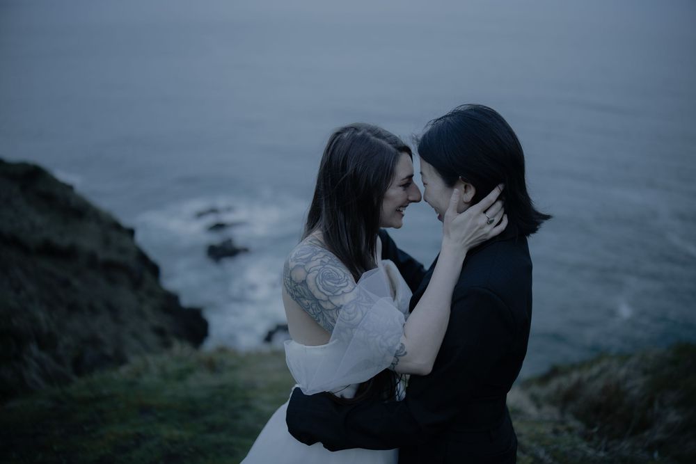 Two brides share a tender moment on a coastal cliff at dusk, one in white lace and one in black, with ocean waves behind them.