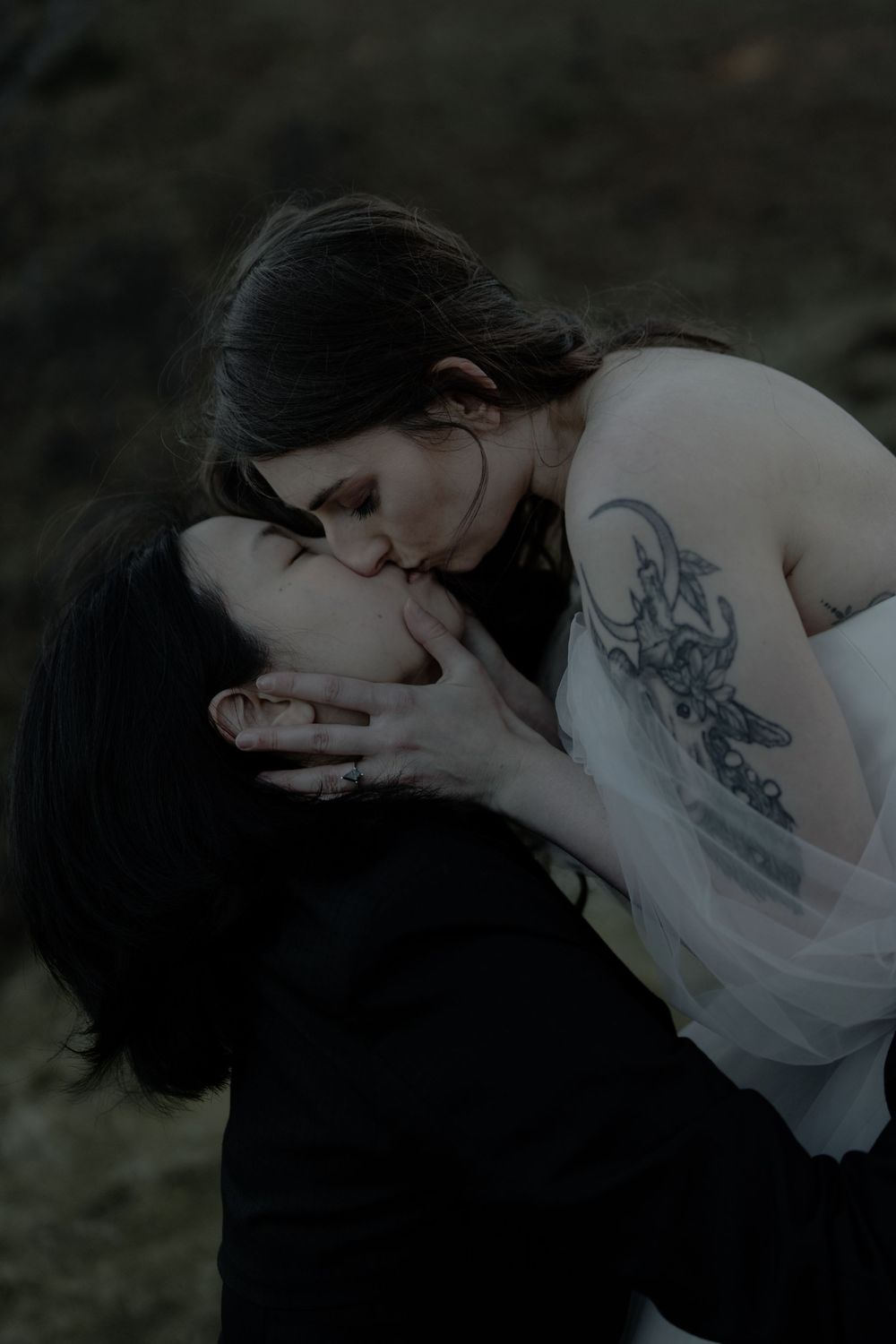 Two women sharing a romantic kiss outdoors, one in white dress with tattoo, other in black, in a dark moody setting.