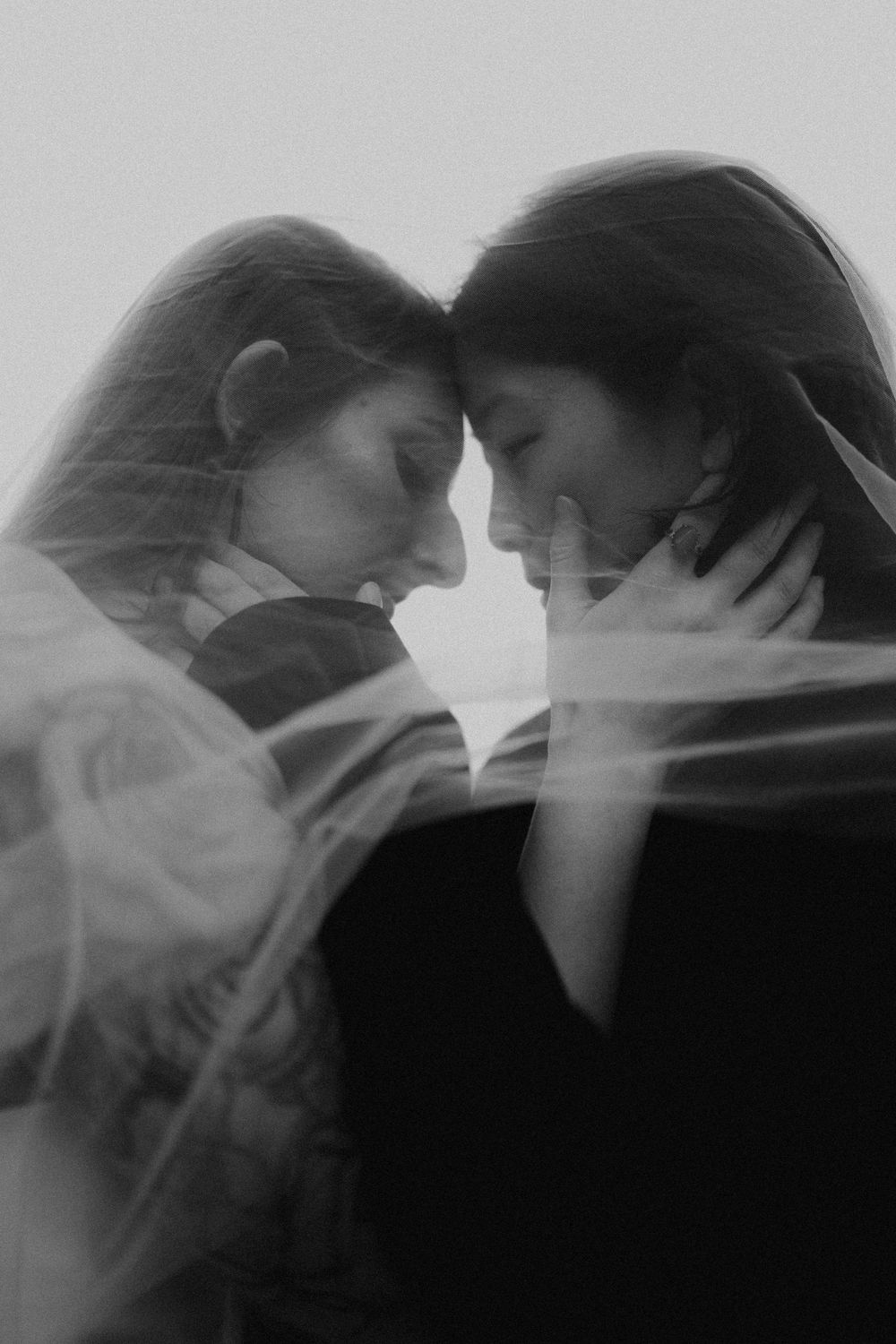 Black and white photo of two women tenderly touching foreheads, draped in a flowing veil, in an intimate embrace.