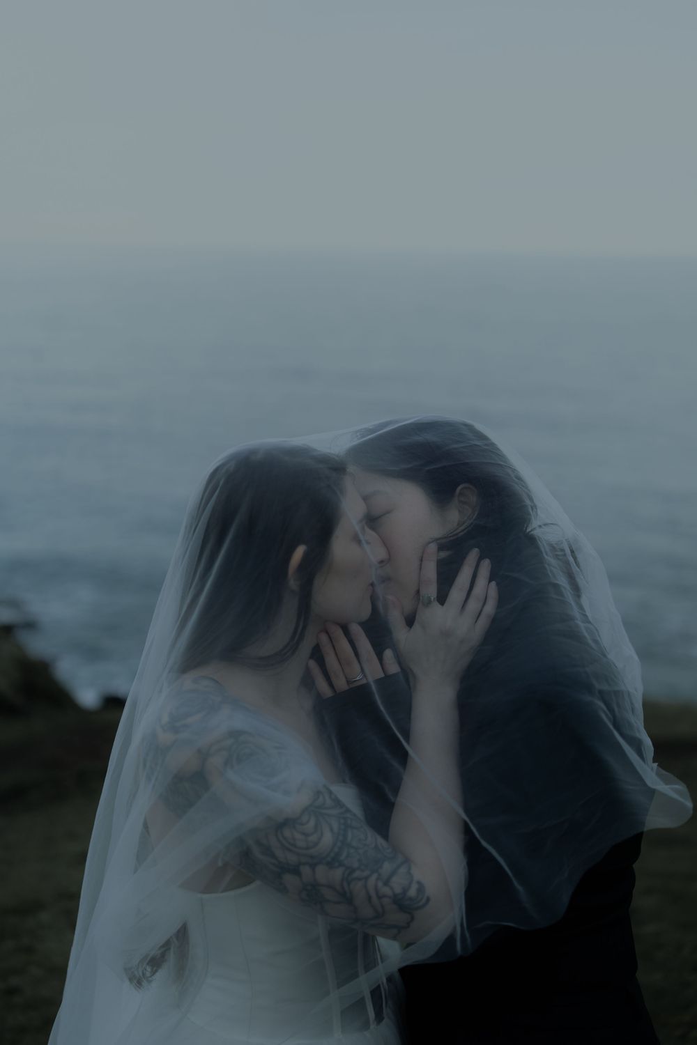 Two brides sharing a kiss under a wedding veil by the ocean at dusk, one with floral tattoos.